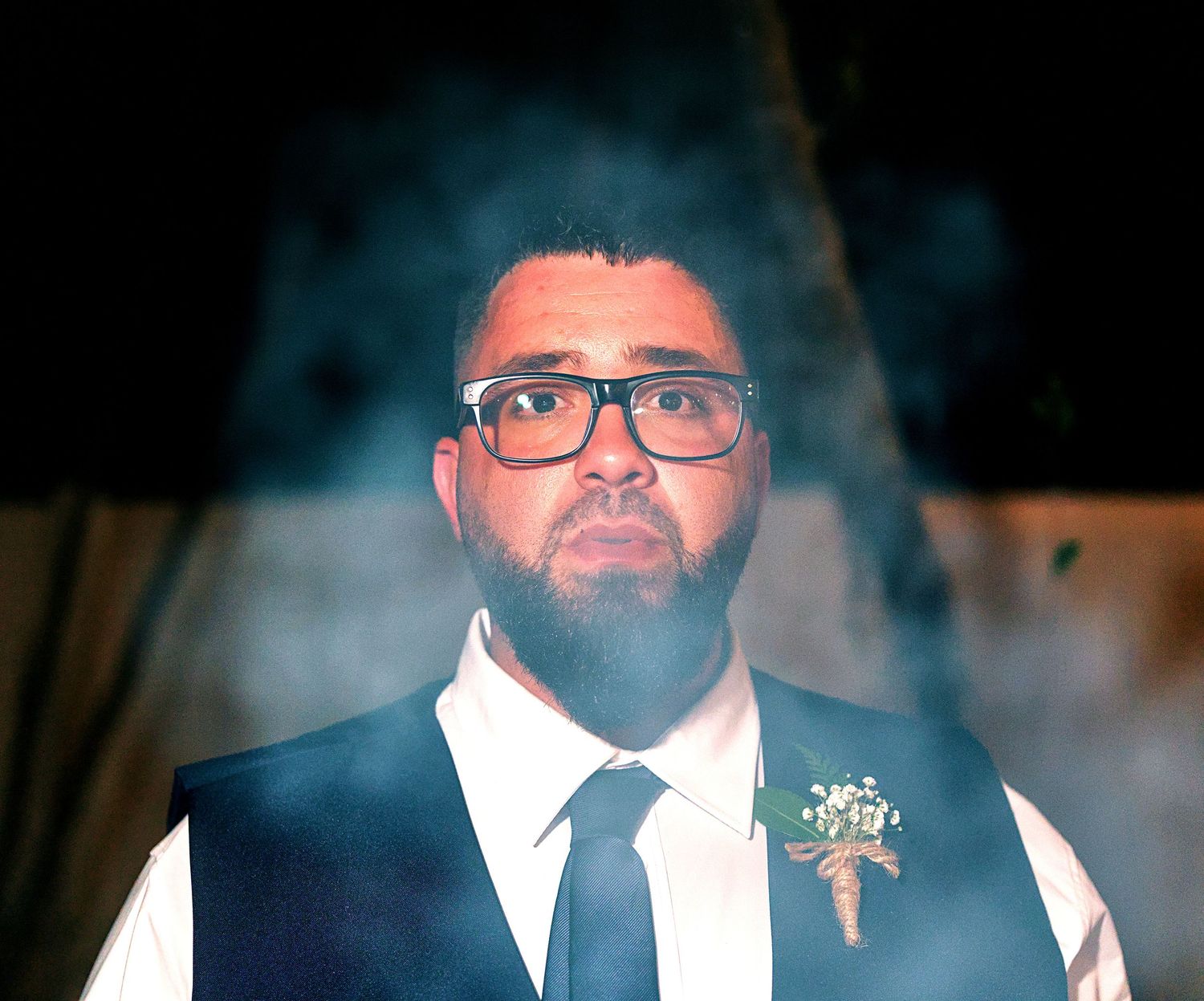Bearded groom in navy vest and tie with boutonniere posed against dramatic smoky background.