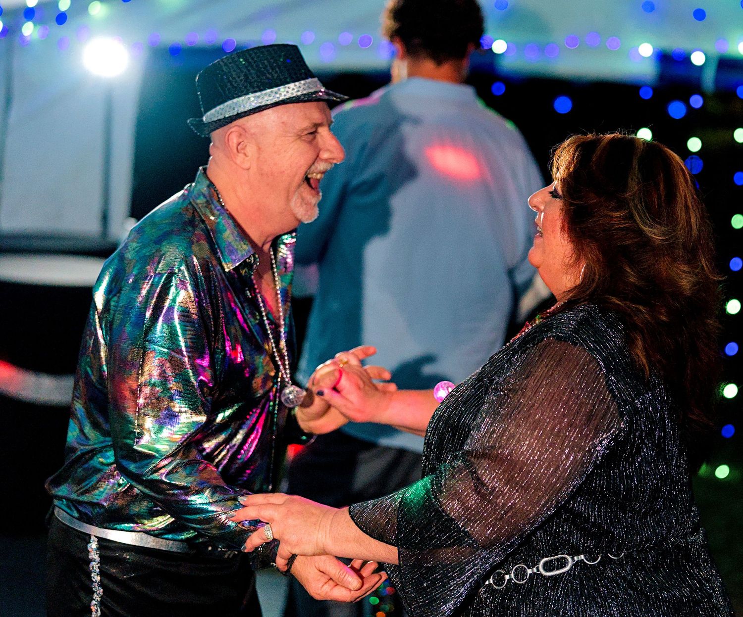 Couple dancing joyfully at a disco-themed party, man wearing a colorful sequin jacket and hat.