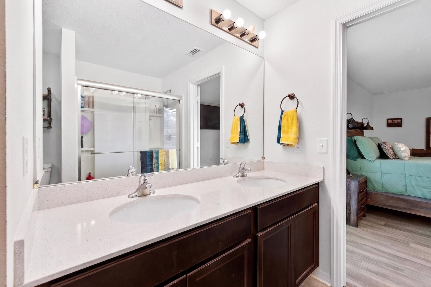 Modern bathroom with double dark wood vanity, white countertops, large mirror, and open view to bedroom.