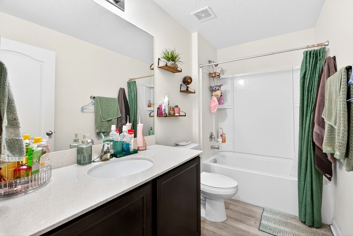 Modern bathroom with dark vanity, white quartz countertop, green shower curtain, and wood-look flooring.