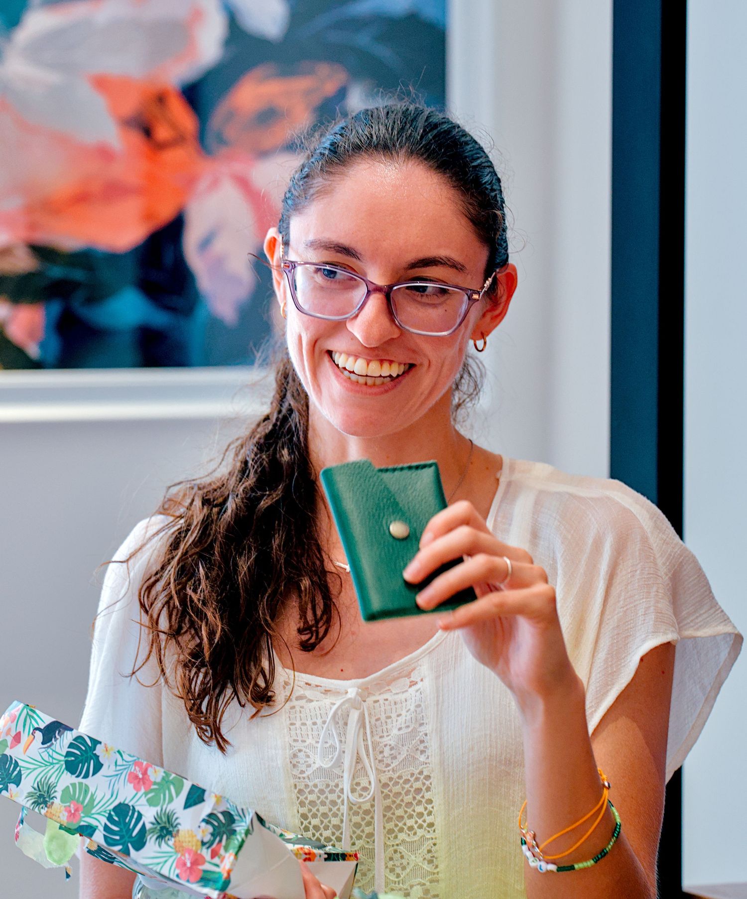 Smiling woman with glasses holding a small green leather wallet, wearing a white top and bracelets, with floral art in background.