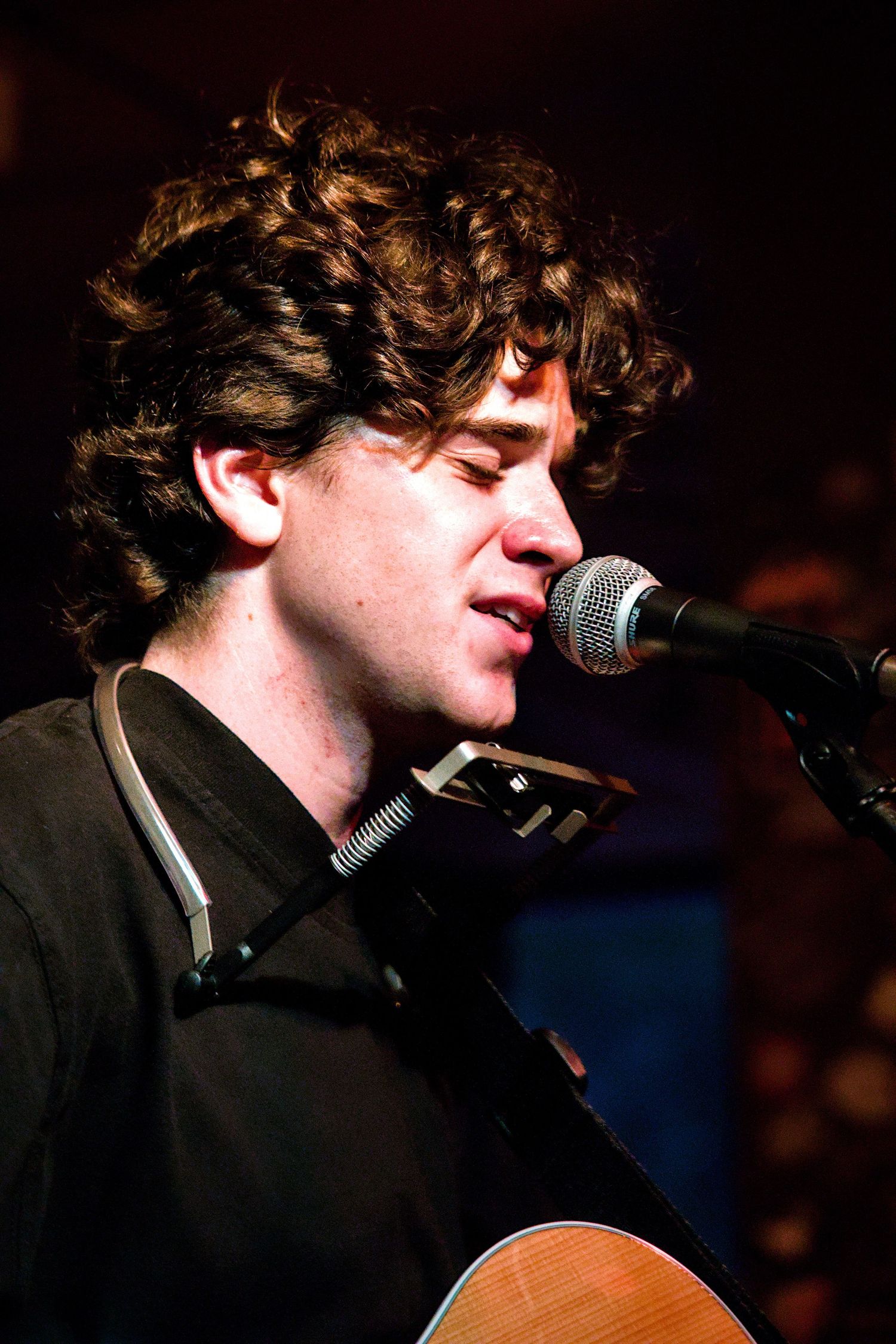 Young curly-haired male musician singing into a microphone while playing acoustic guitar on stage.