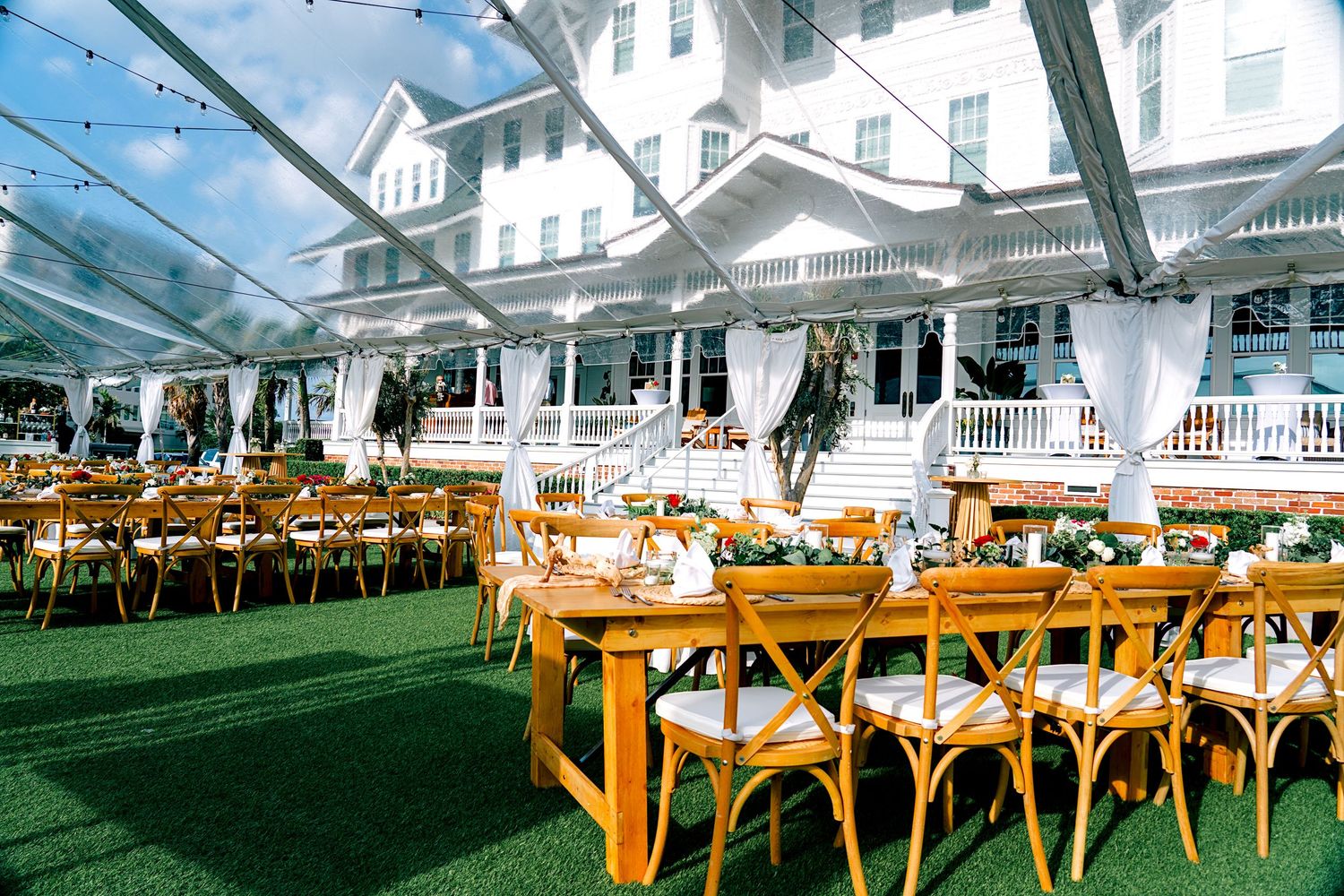 Elegant outdoor wedding reception under a clear tent with wooden cross-back chairs and long decorated tables on green lawn.