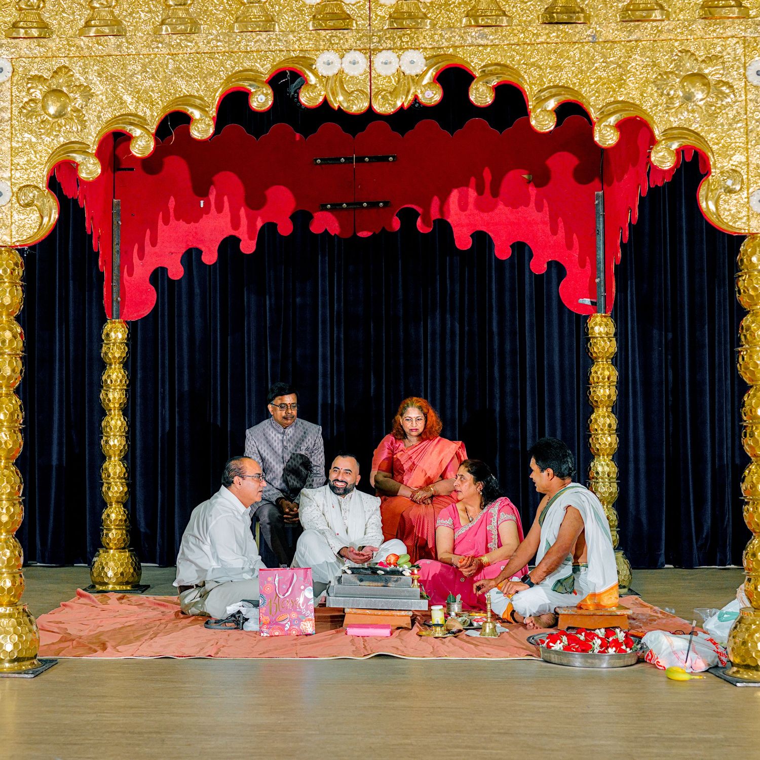 Group performing Hindu puja ceremony seated on floor before ornate golden temple altar with red canopy.