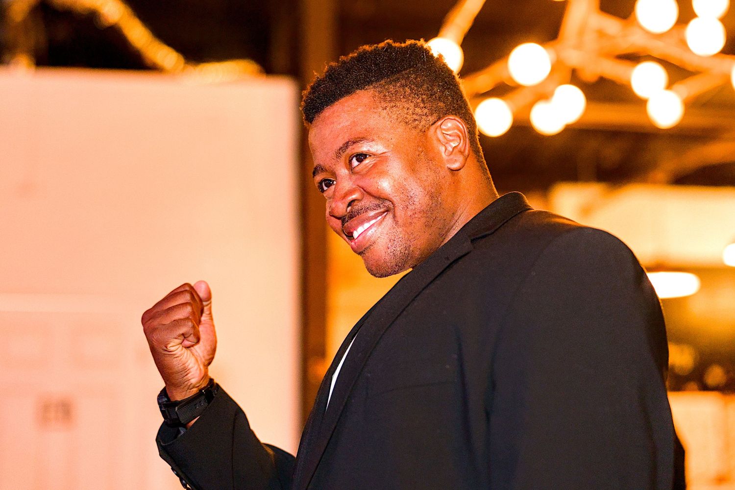 A smiling man in a black blazer raises his fist triumphantly at an evening event with warm bokeh lights.