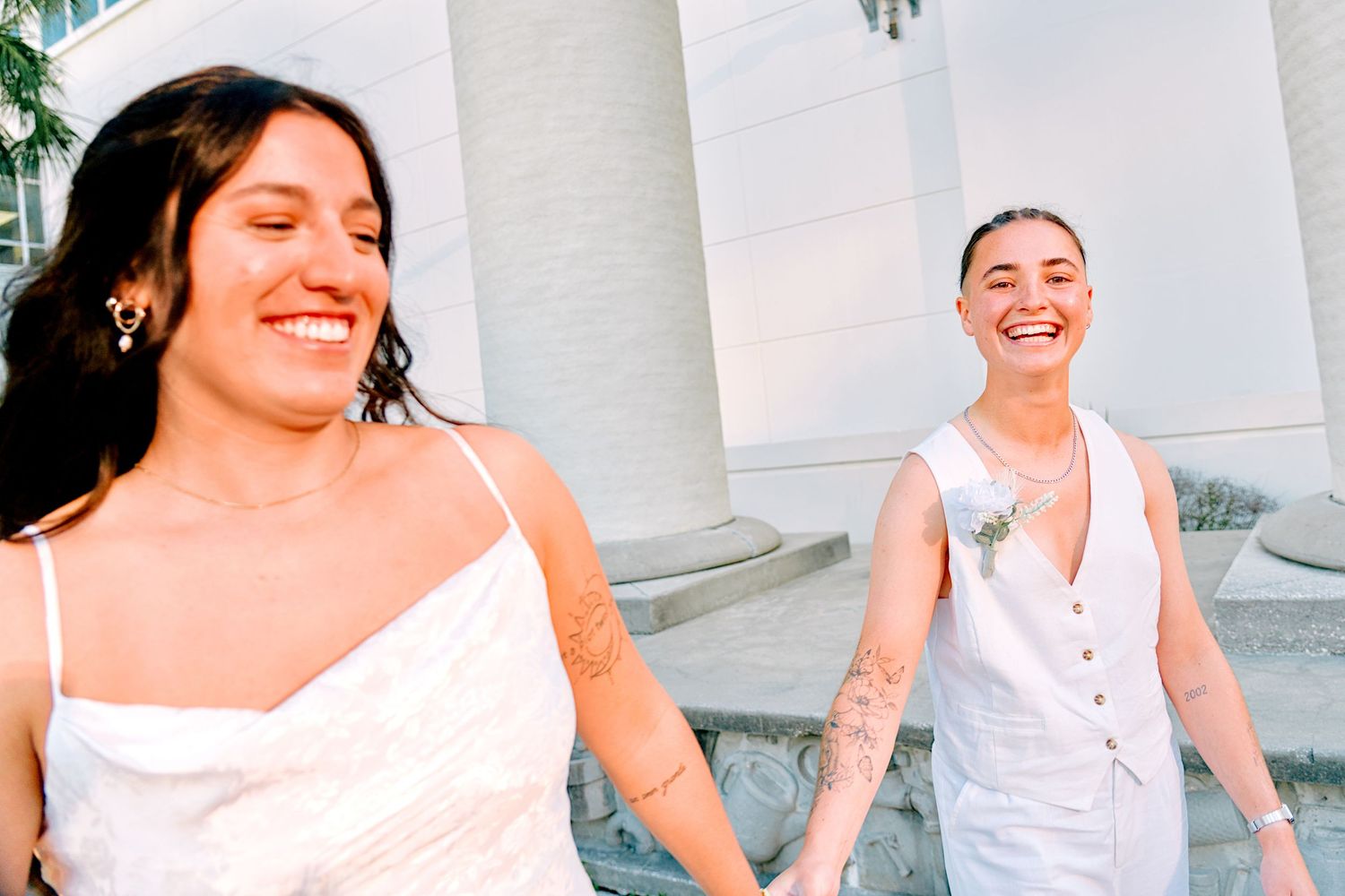 Two smiling brides in white wedding attire hold hands outdoors near white columns on their wedding day.