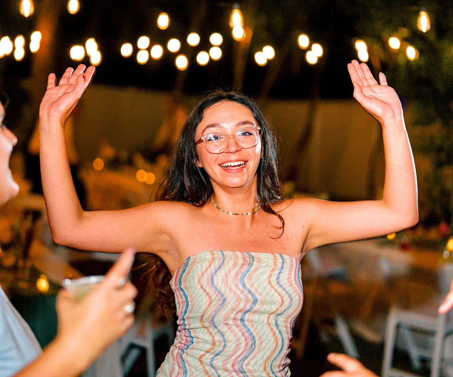 Young woman in colorful strapless dress dancing joyfully with hands raised at an outdoor evening wedding reception.