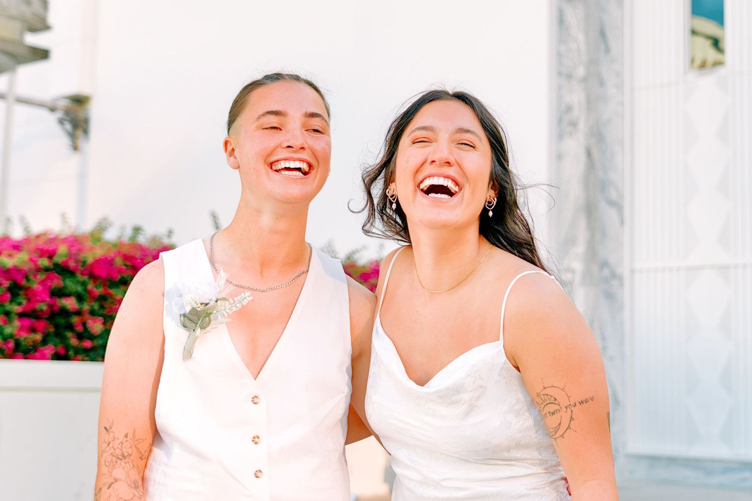 Two brides laughing joyfully on their wedding day, wearing white dresses with floral details and tattoos visible.