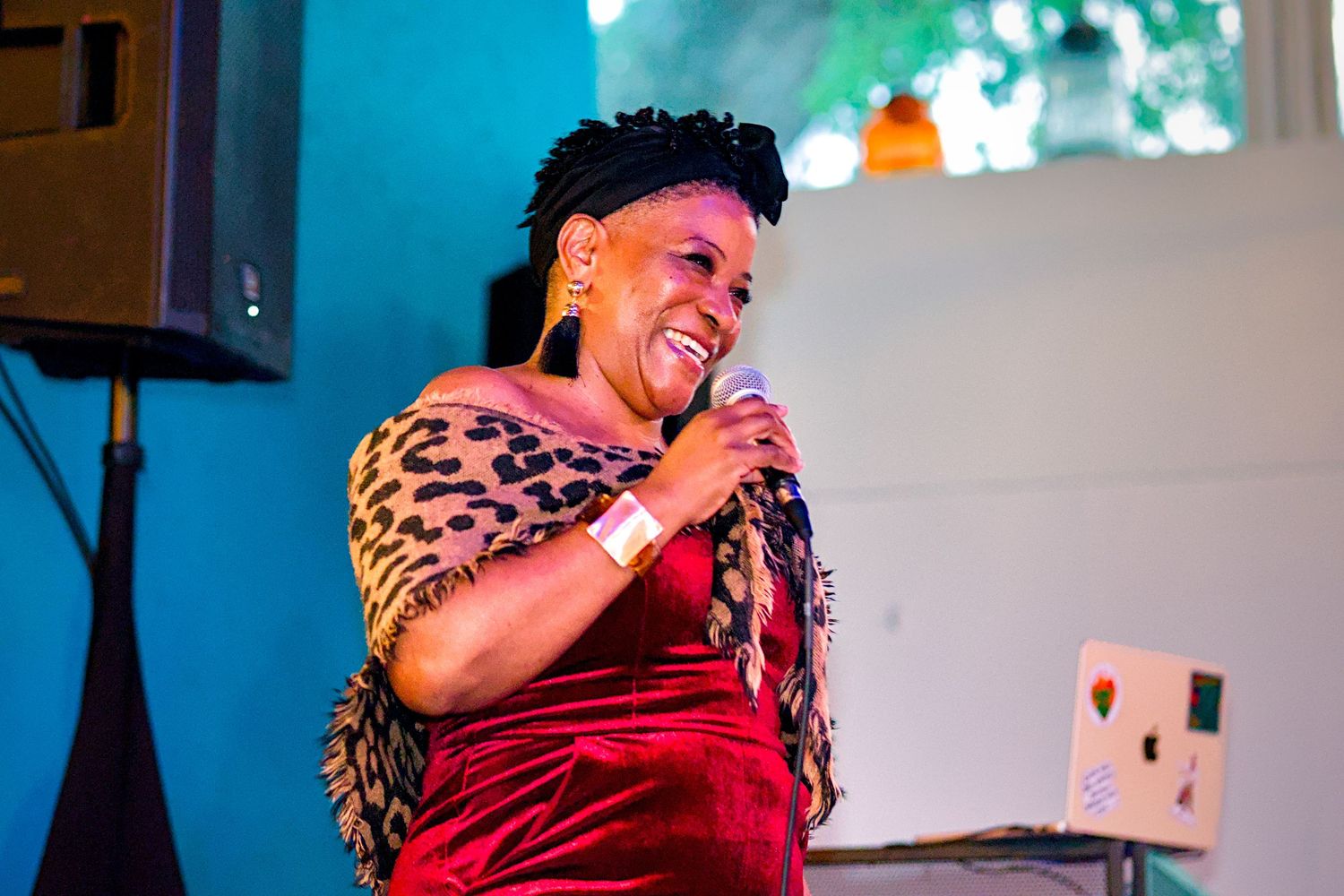 Woman in leopard print and red velvet singing into microphone on stage at an indoor event.