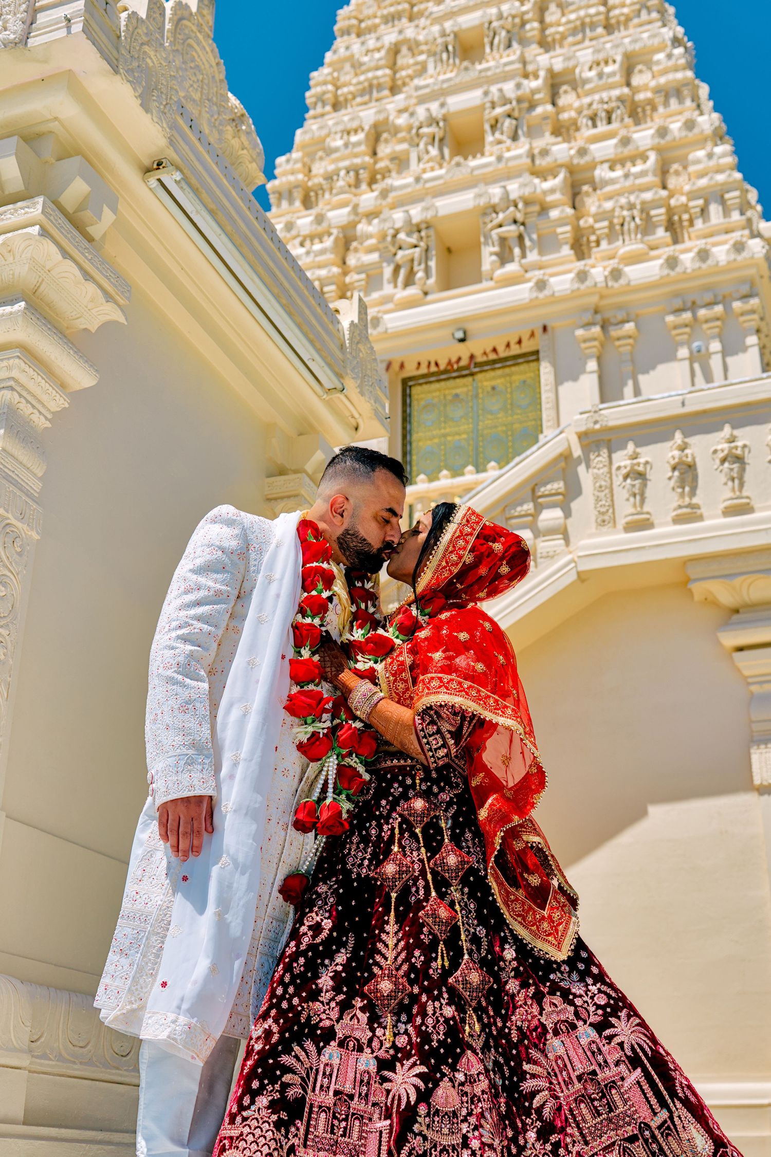 Indian bride in red lehenga and groom in white sherwani share a romantic moment outside a ornate Hindu temple.