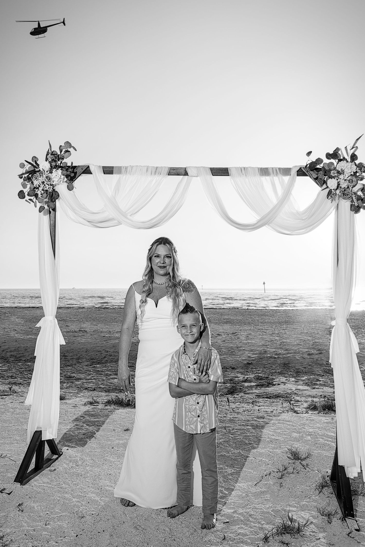Black and white photo of bride in white gown posing with young boy under decorated beach wedding arch.