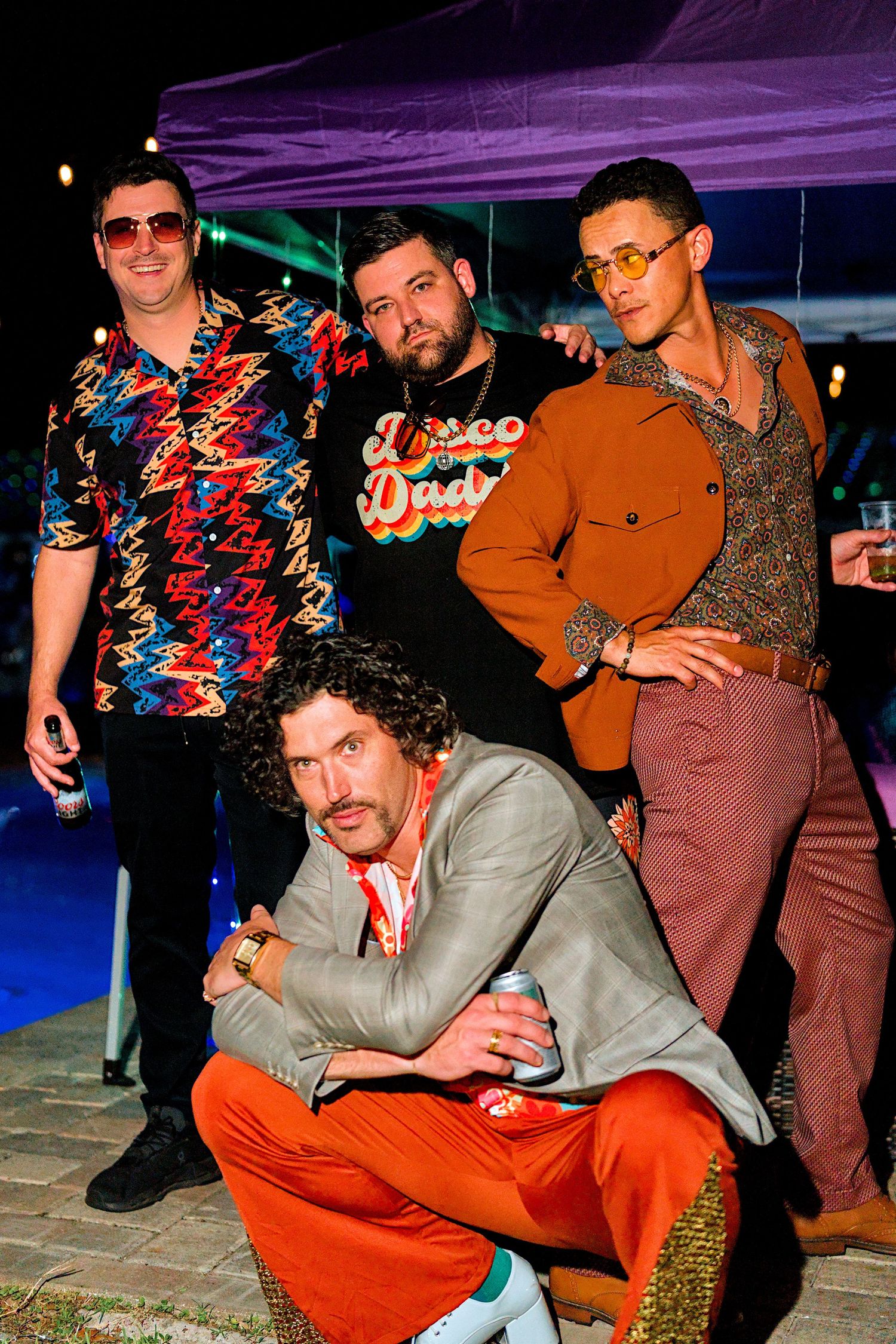 Four men in 1970s disco-themed outfits posing together at an outdoor retro party event at night.