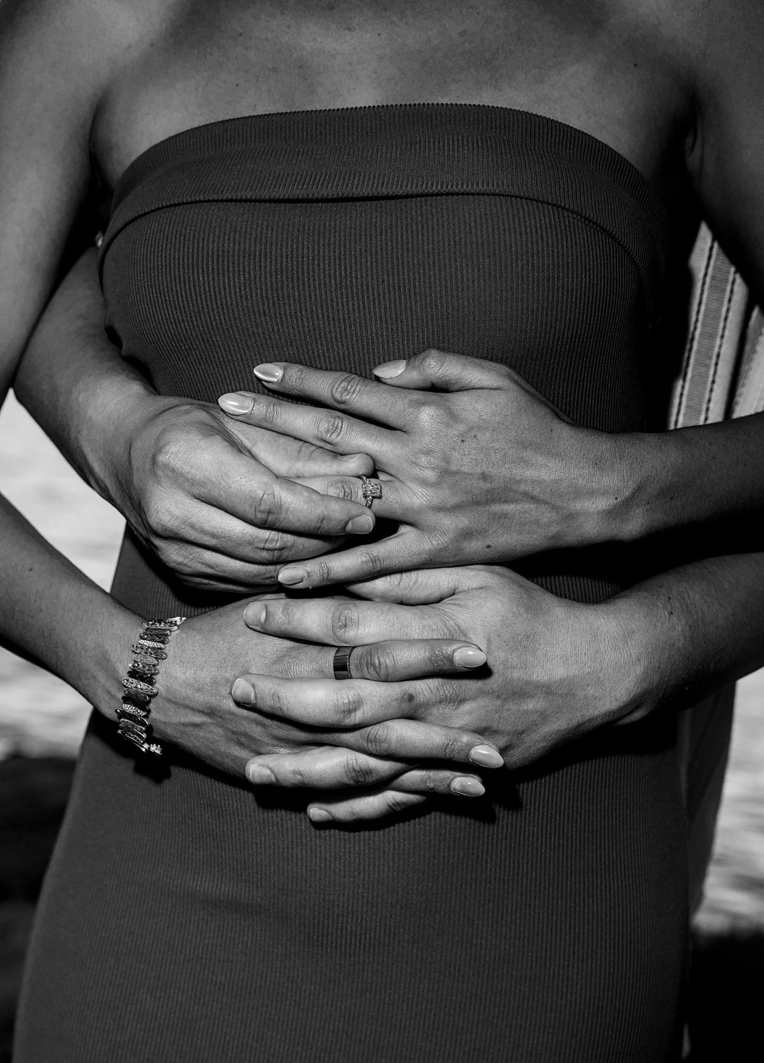 Black and white photo of two people embracing, hands clasped over a pregnant belly, wearing wedding rings.