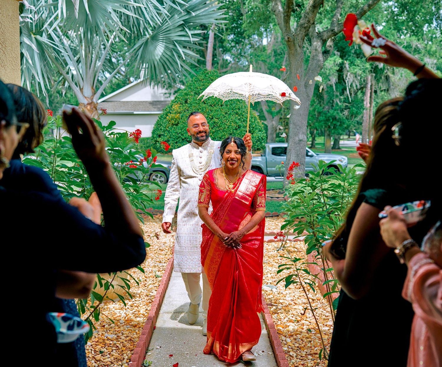 Indian wedding couple walking under white umbrella through floral petal toss celebration.