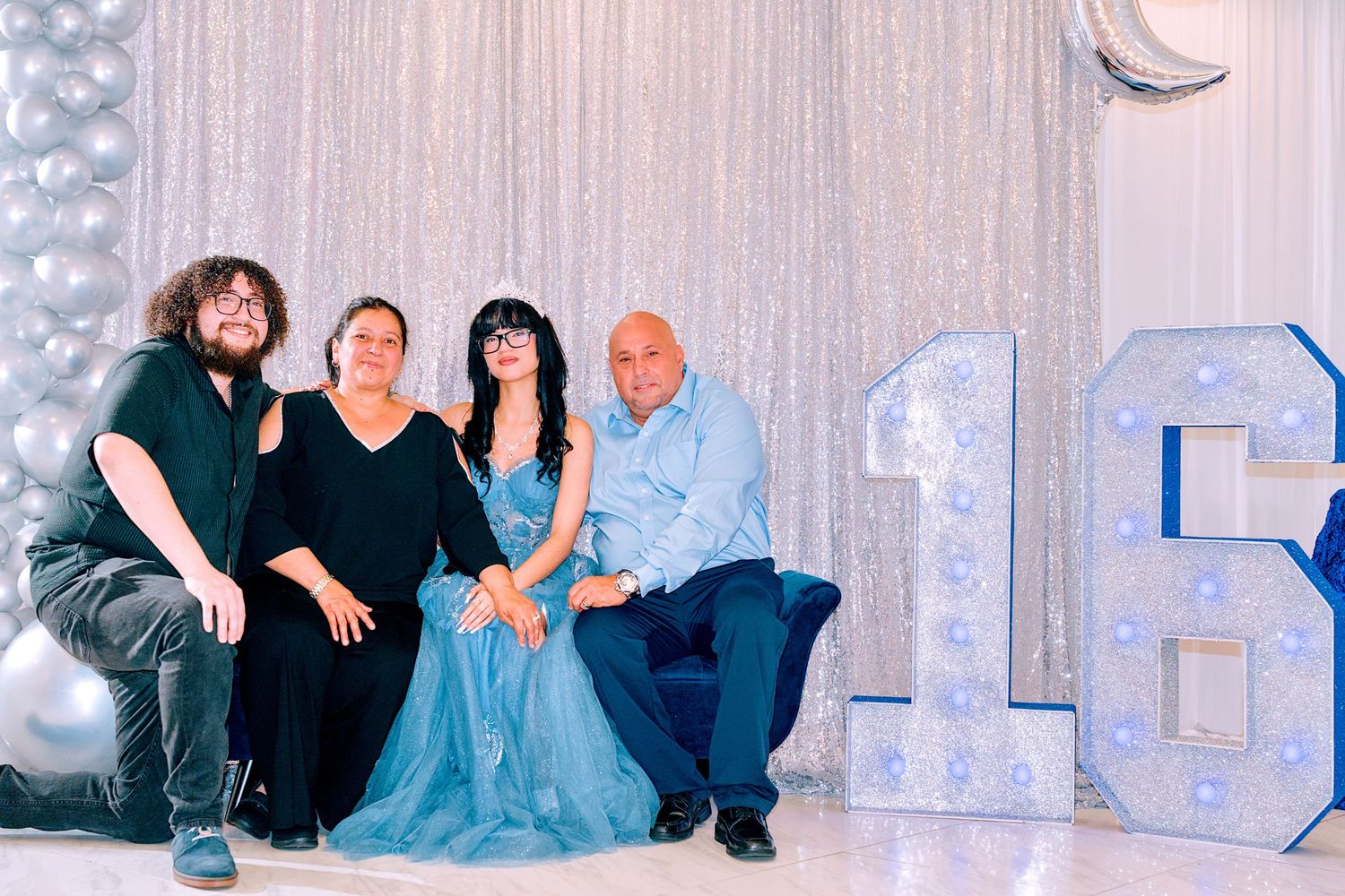 Four people pose elegantly at a quinceañera celebration beside illuminated number 16 decorations and silver balloons.