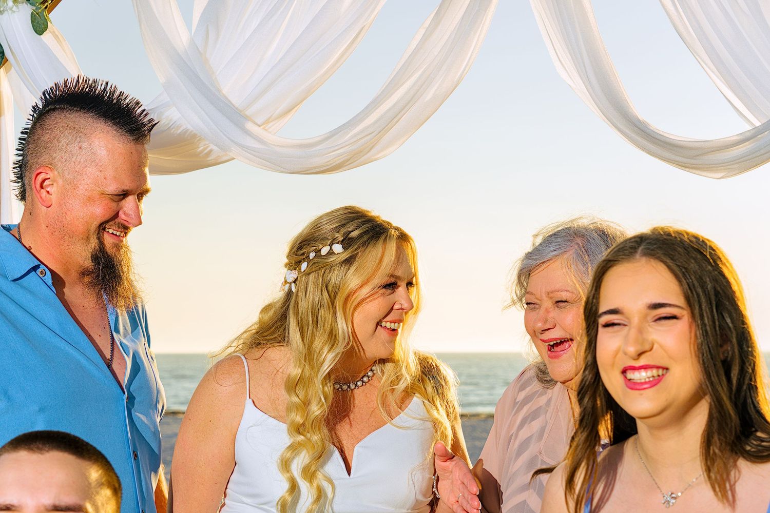 Wedding party gathered under white fabric canopy at beach during sunset celebration.