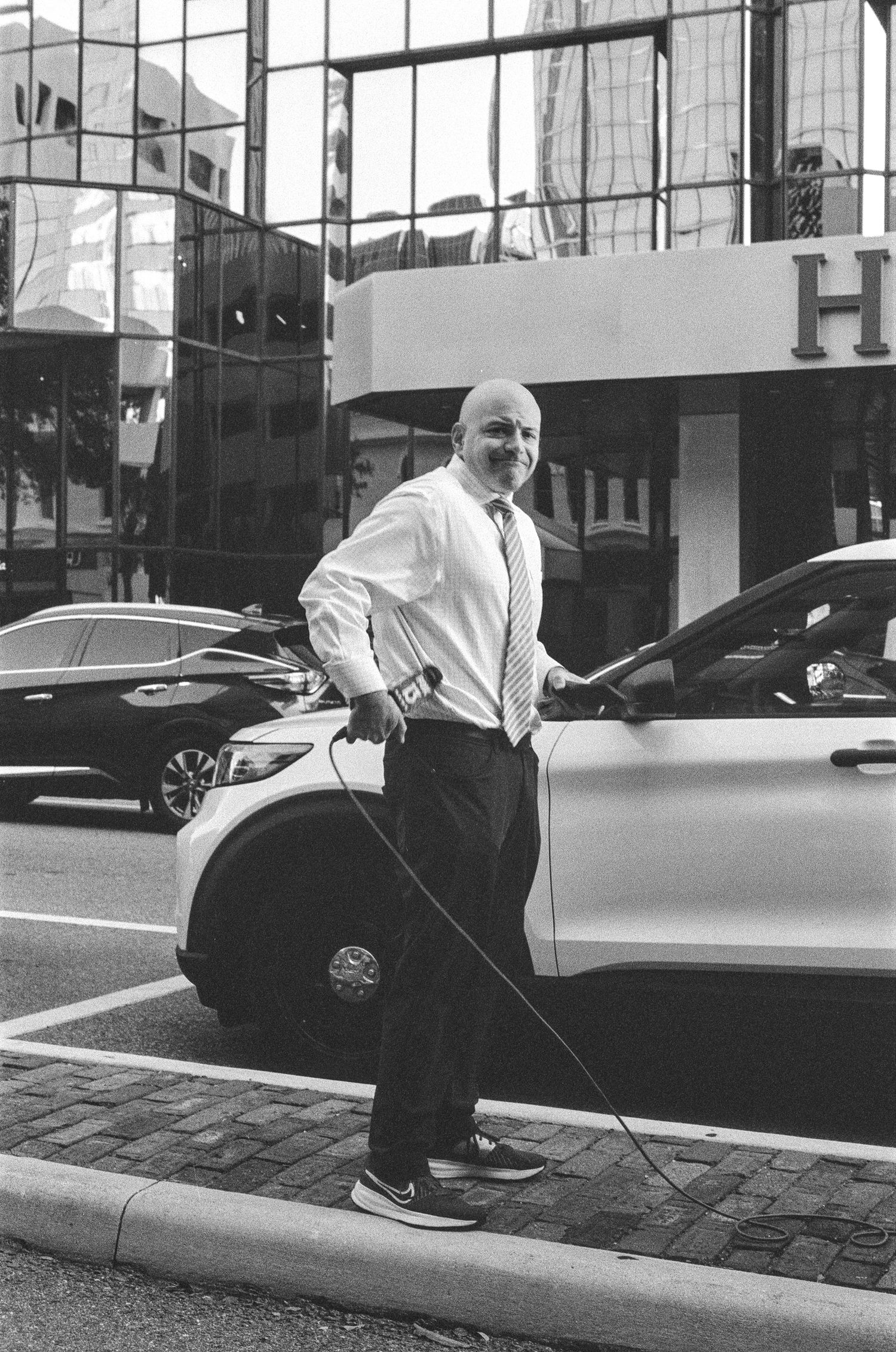 Bald man in dress shirt and tie charging an electric car outside a modern glass building in black and white.