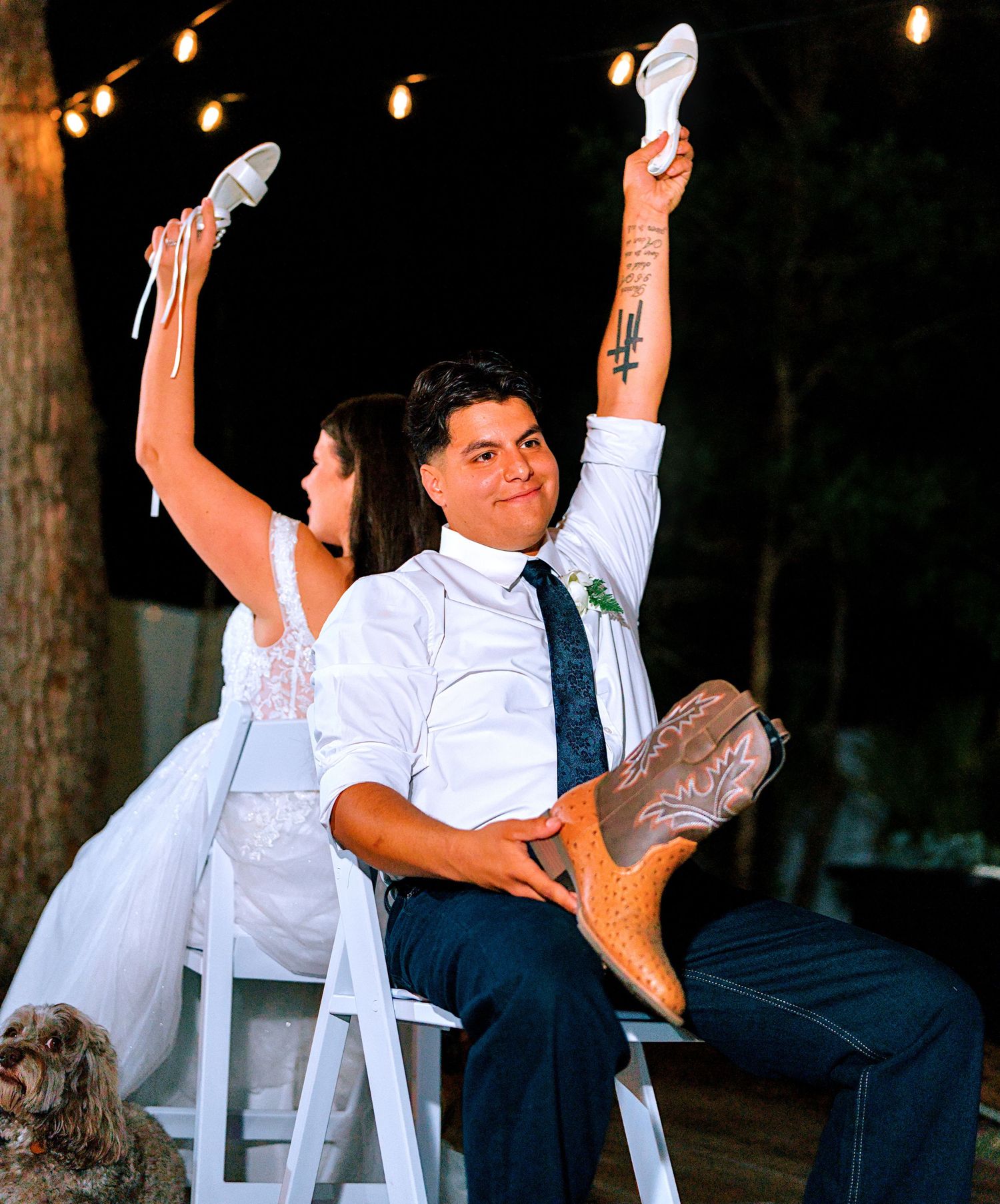 Bride and groom holding up shoes during wedding shoe game while seated in white chairs outdoors at night.