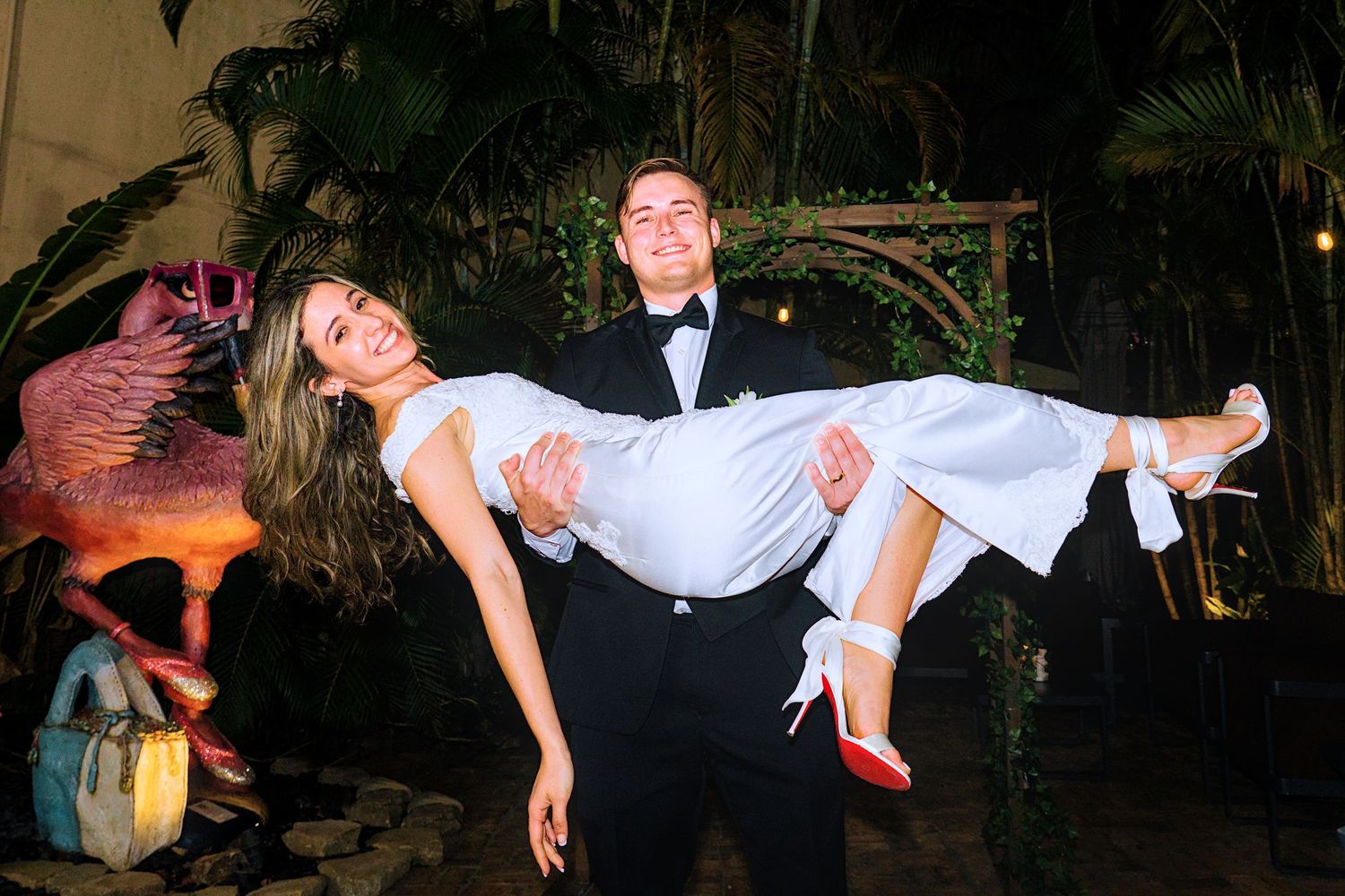 Groom in tuxedo carries bride in white wedding dress outdoors near tropical plants and flamingo statue.