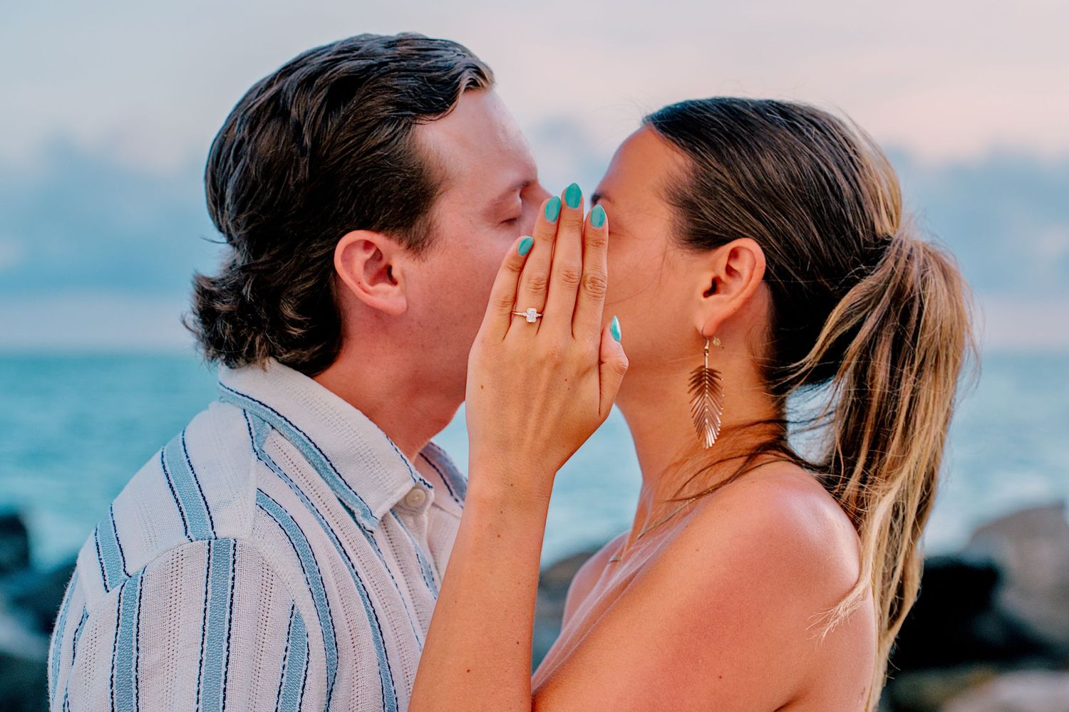 Couple shares romantic kiss on beach at sunset after engagement, woman showing ring on hand.
