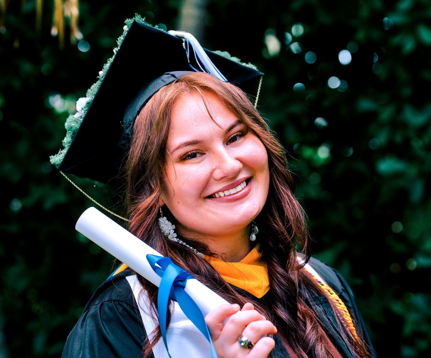 Smiling young woman in black graduation cap and gown holding diploma outdoors.