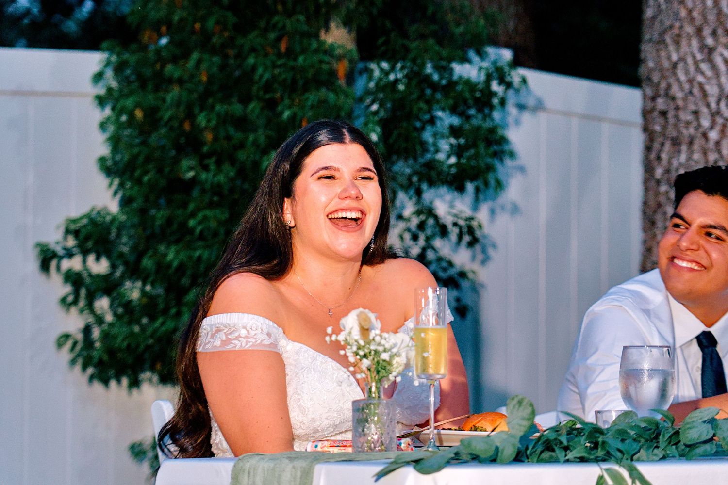 Laughing bride in off-shoulder lace gown seated at sweetheart table during wedding reception.