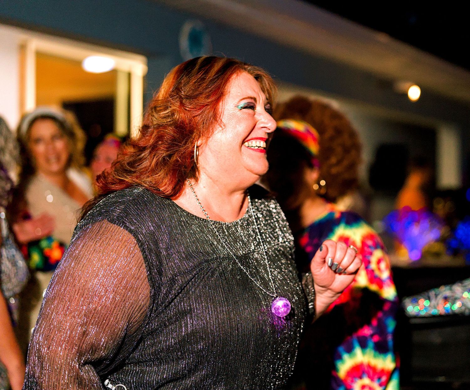 A joyful woman with red hair dancing and laughing at a colorful party event wearing a sparkly dark outfit.