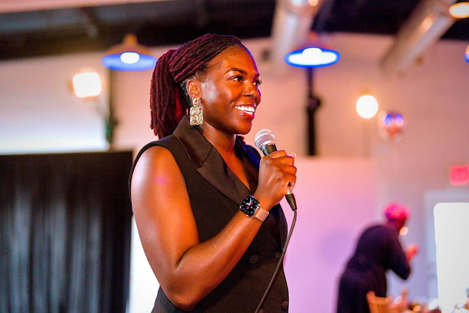 A woman in a black sleeveless dress smiles while speaking into a microphone at an indoor event.