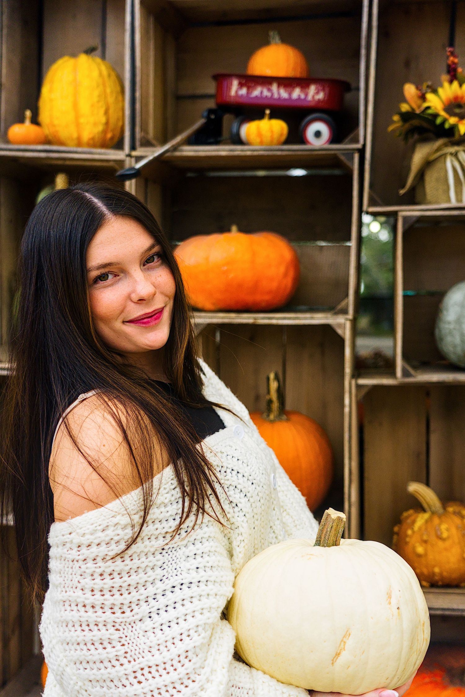 Woman in white sweater poses with autumn pumpkins and gourds in rustic wooden display shelves.
