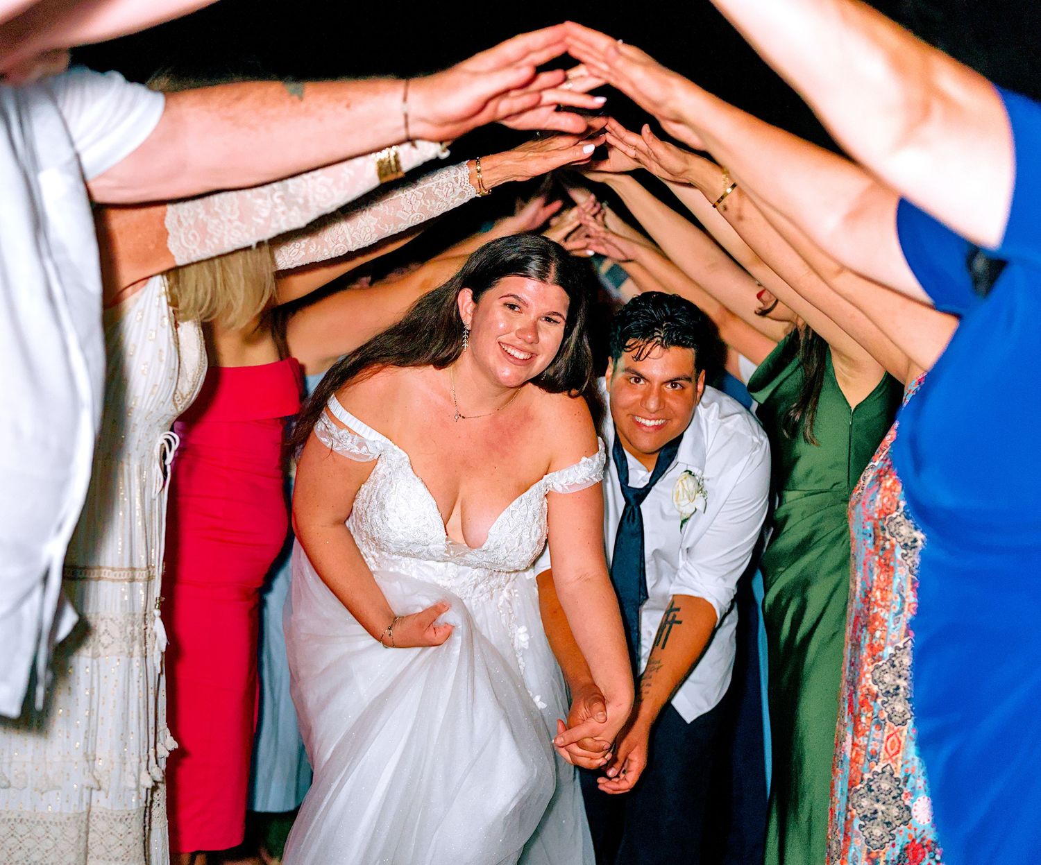Bride and groom walk hand-in-hand through tunnel of raised hands formed by wedding guests at reception.