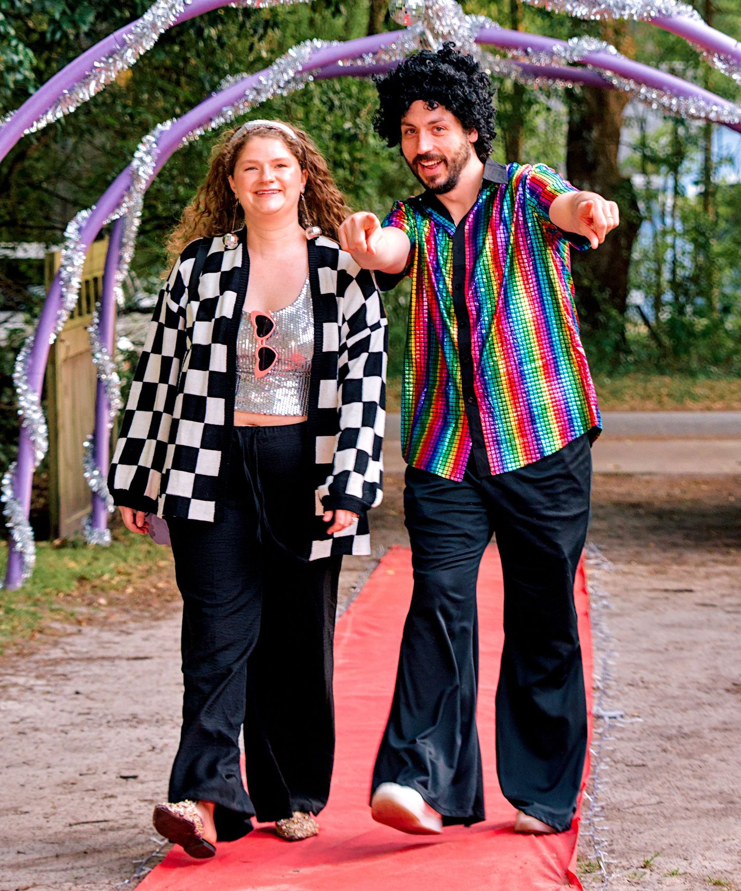 Couple in disco costumes posing on red carpet outdoors at themed event.