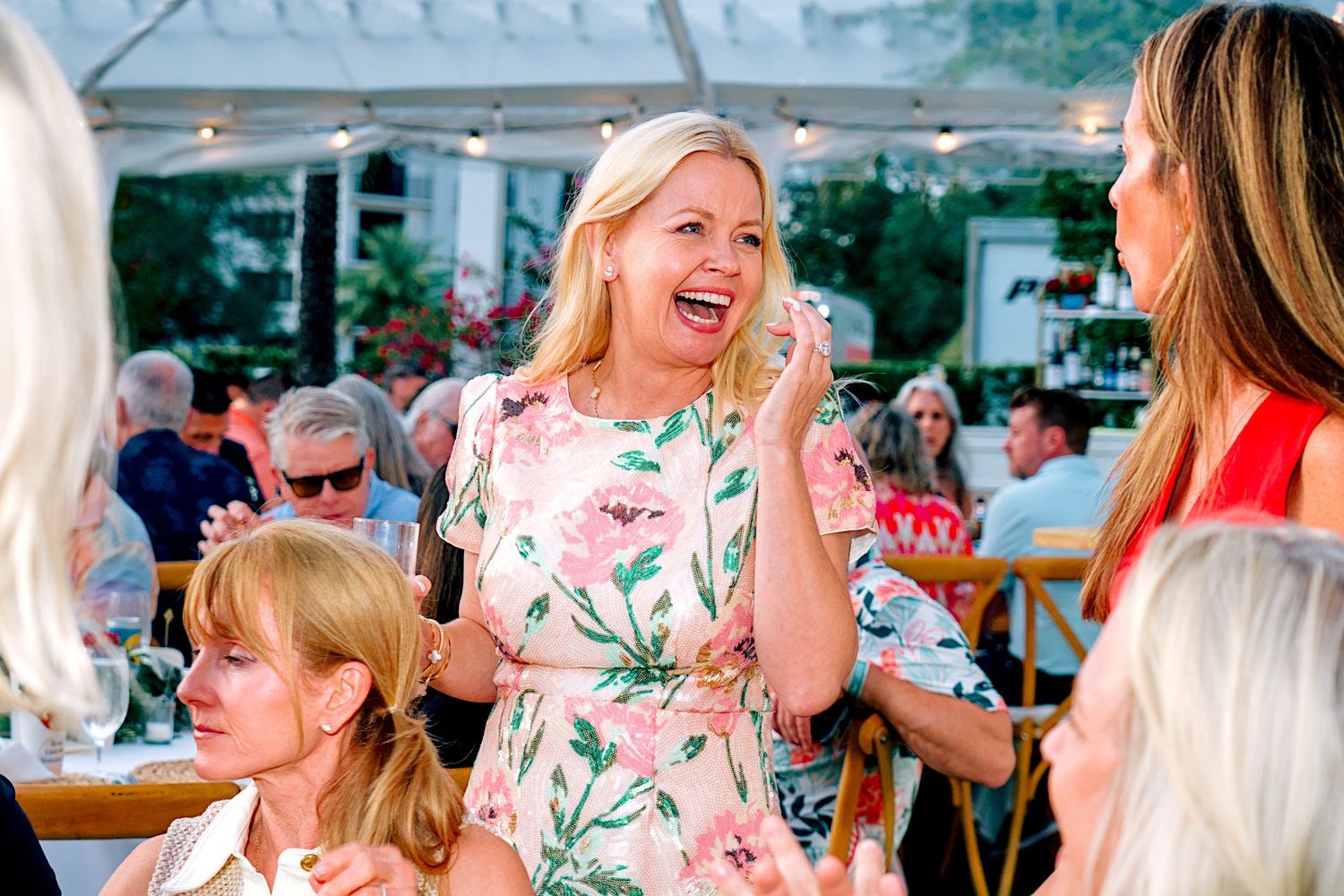 Joyful blonde woman in floral dress laughing at an outdoor social gathering event with guests in background.