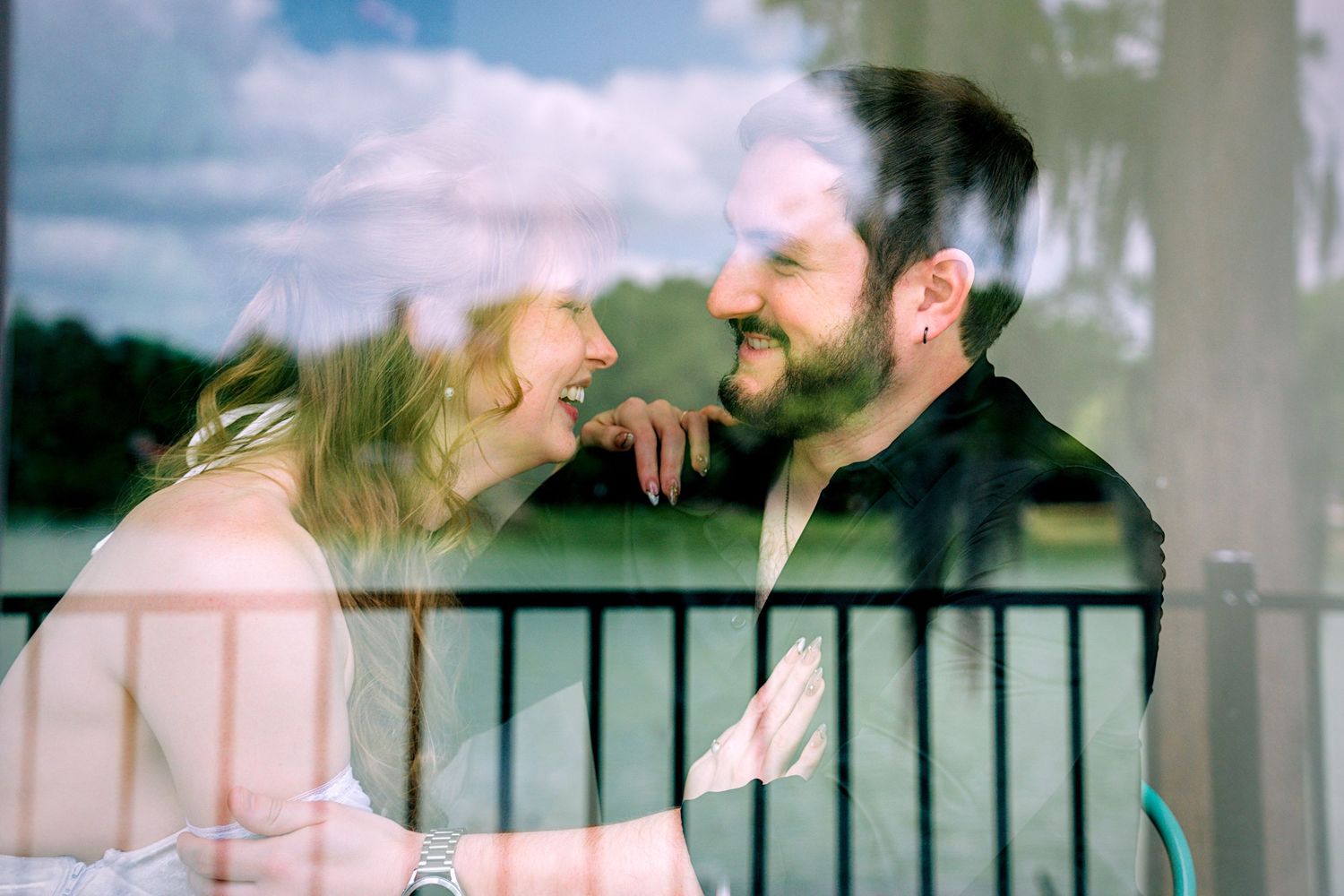 Couple smiling at each other through a glass window with outdoor reflection, captured in a romantic candid moment.