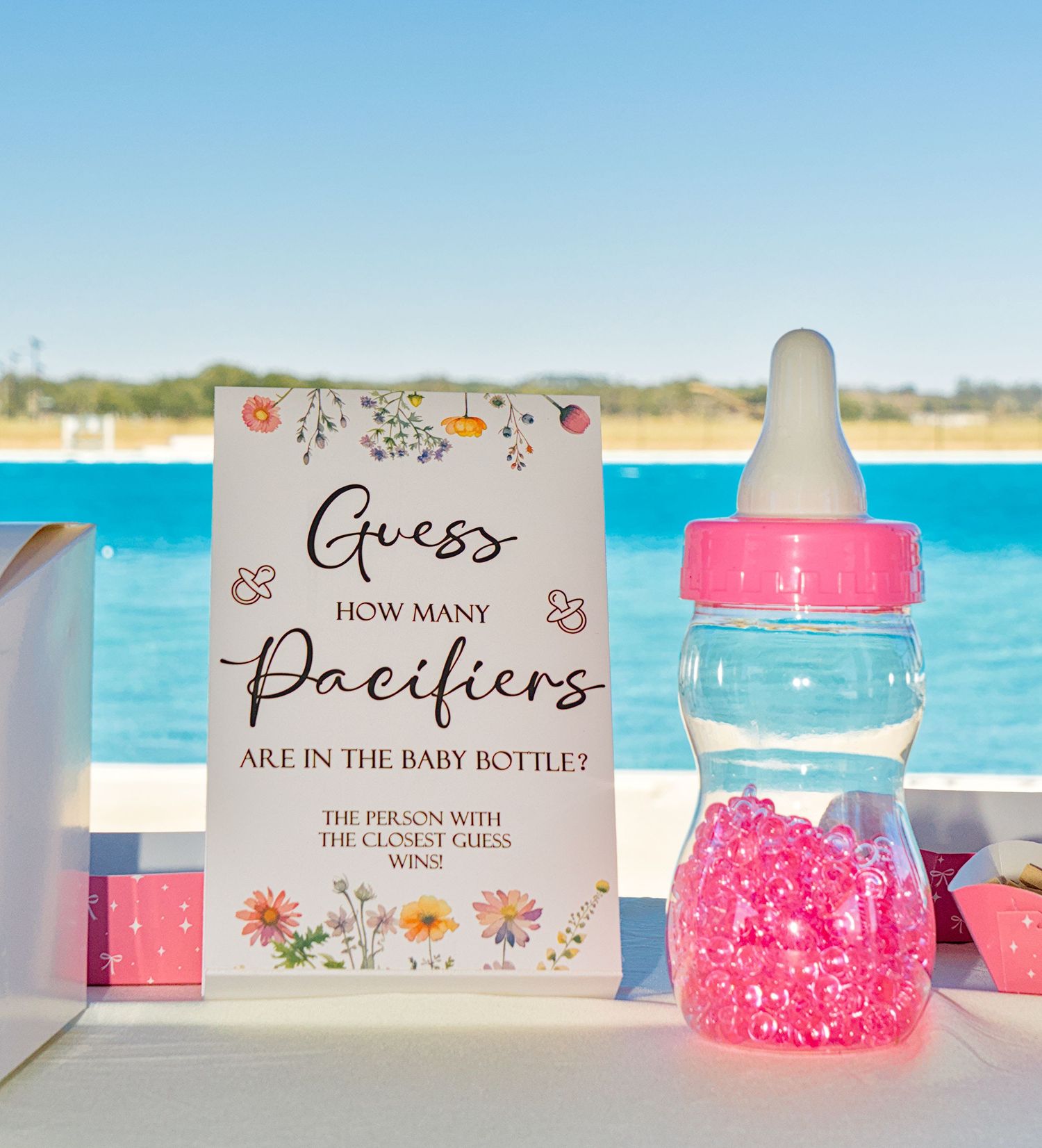 Baby shower game with pink candy-filled bottle and 'Guess How Many Pacifiers' sign against blue ocean backdrop.