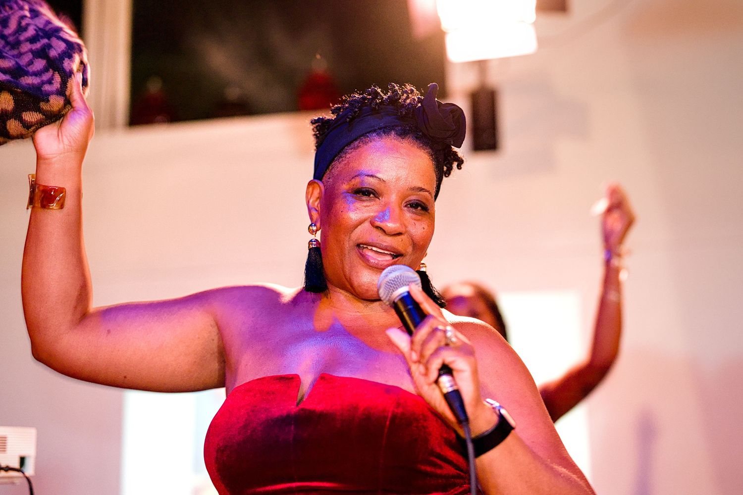 Joyful Black woman in a red velvet dress sings into a microphone while raising her arm at an event.