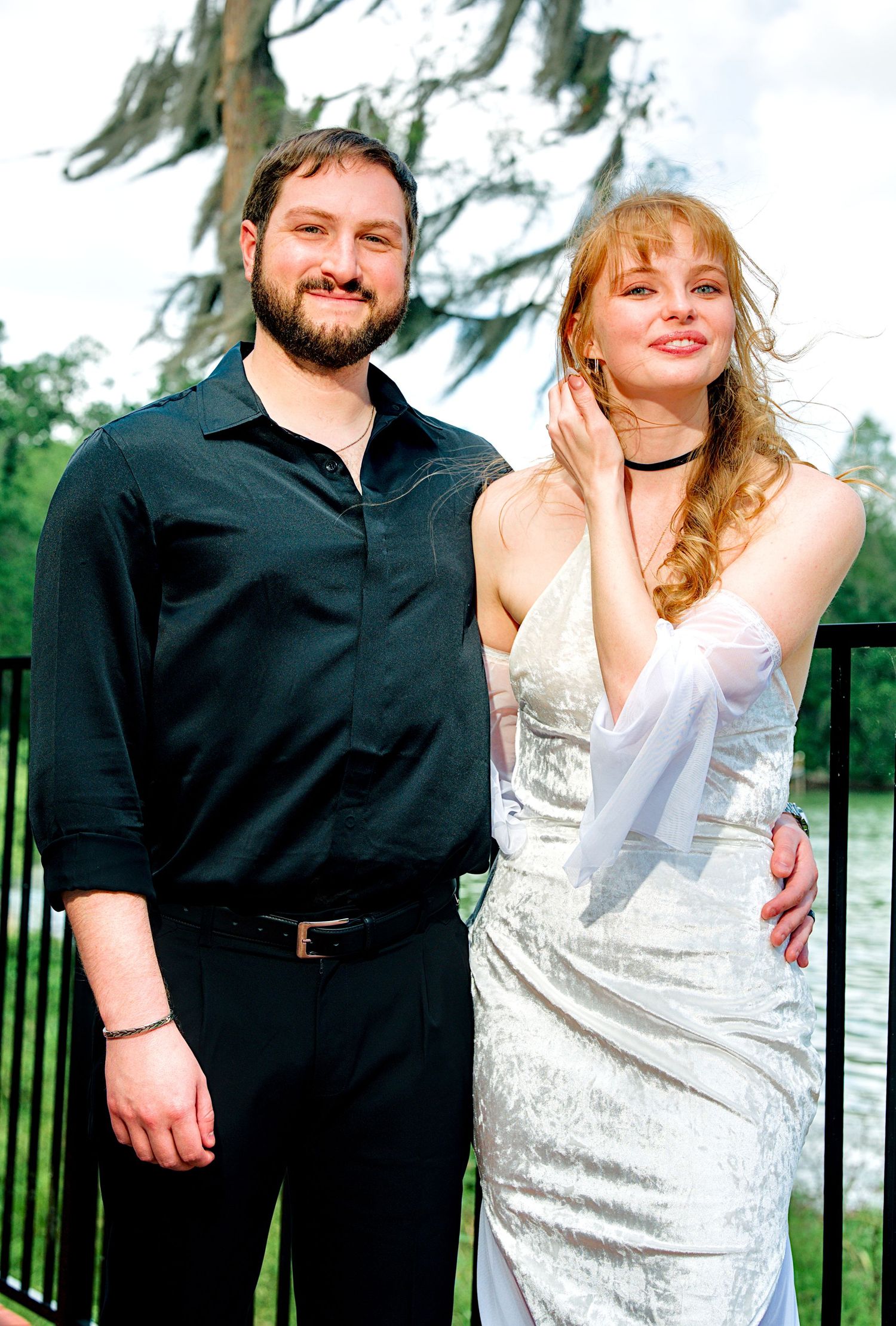 Couple posing outdoors near water, man in black shirt with arm around woman in white dress.