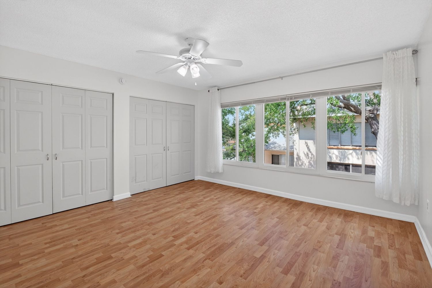 Empty white room with hardwood flooring, ceiling fan, and large windows letting in natural light.