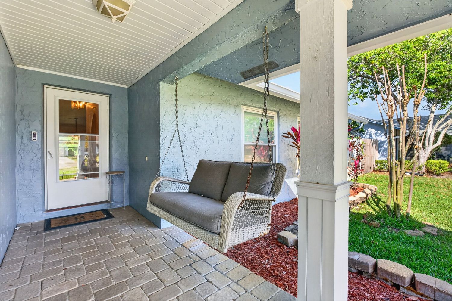Covered front porch with wicker hanging swing, brick pavers, and lush green landscaping on a sunny day.