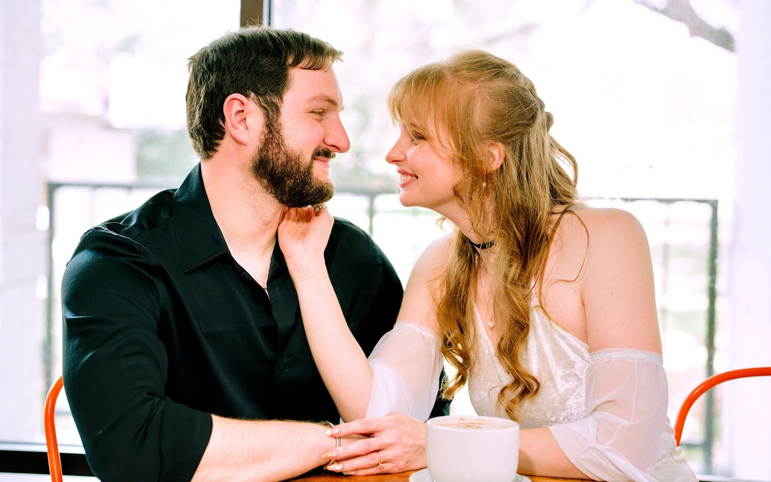 Smiling bride and groom in black jacket sharing a romantic moment at a cafe table.