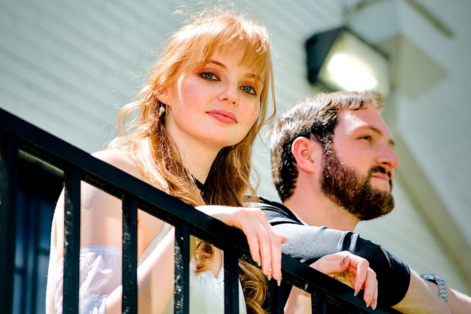 A young woman with long red hair and a bearded man pose together on a balcony in elegant attire.