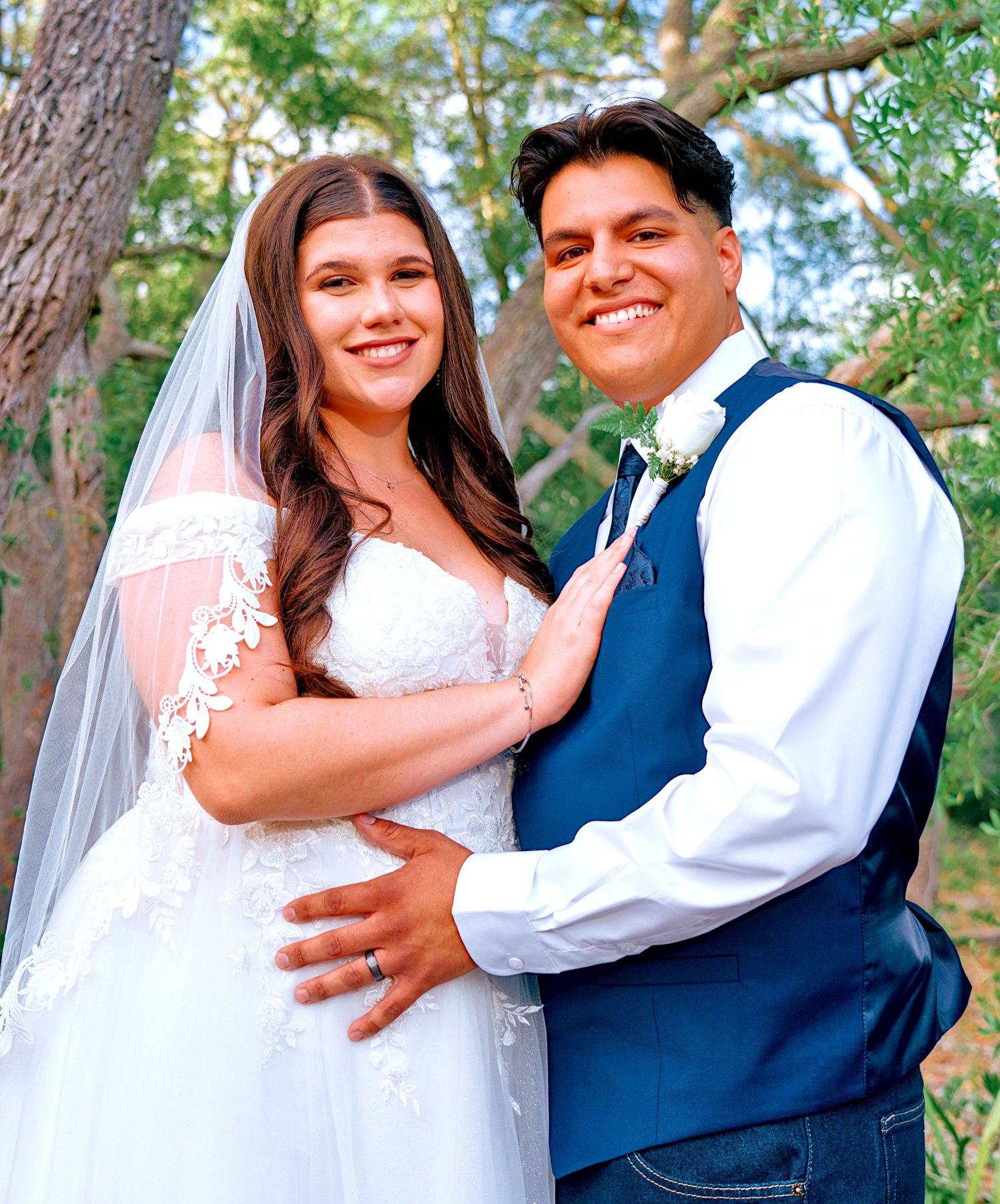 Smiling bride in lace wedding dress and veil poses with groom in blue vest outdoors among trees.