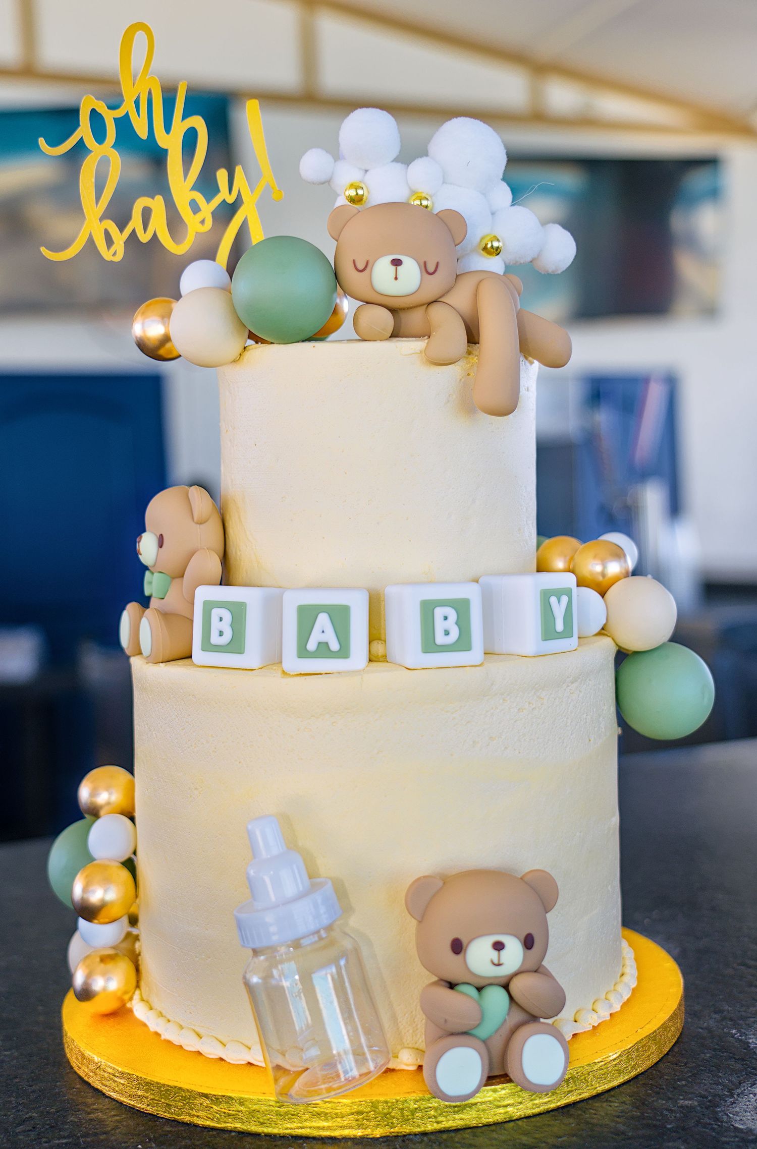 Elegant two-tiered baby shower cake decorated with teddy bears, baby bottles, and gold accents on a yellow cake board.