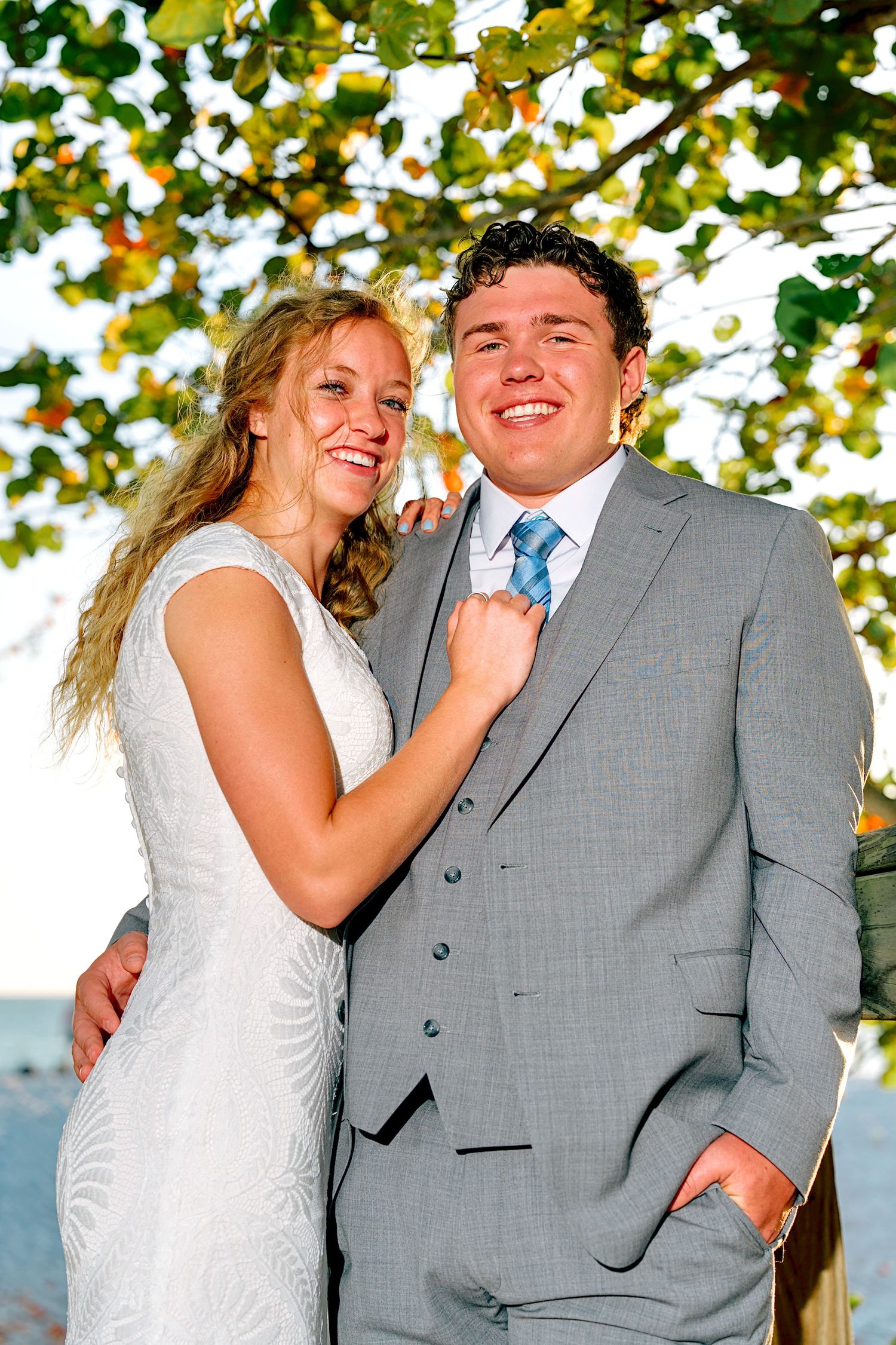 Smiling bride and groom pose outdoors under lush green trees near the water on their wedding day.