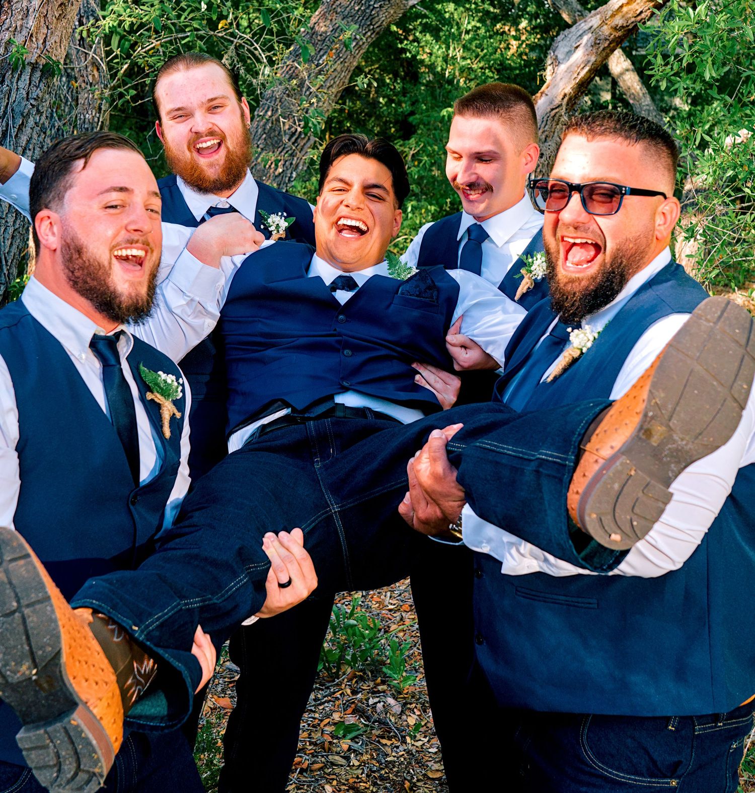 Groomsmen in navy suits laughing while lifting the groom outdoors at a wedding.