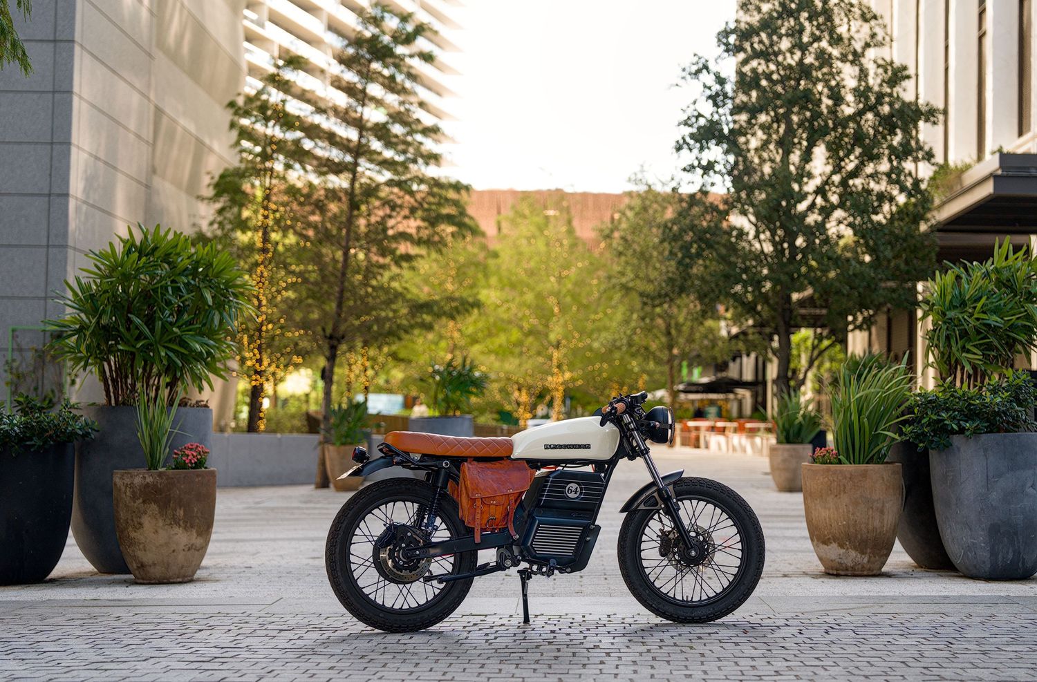 Vintage orange and white motorcycle parked on cobblestone plaza surrounded by lush trees and modern buildings at sunset.