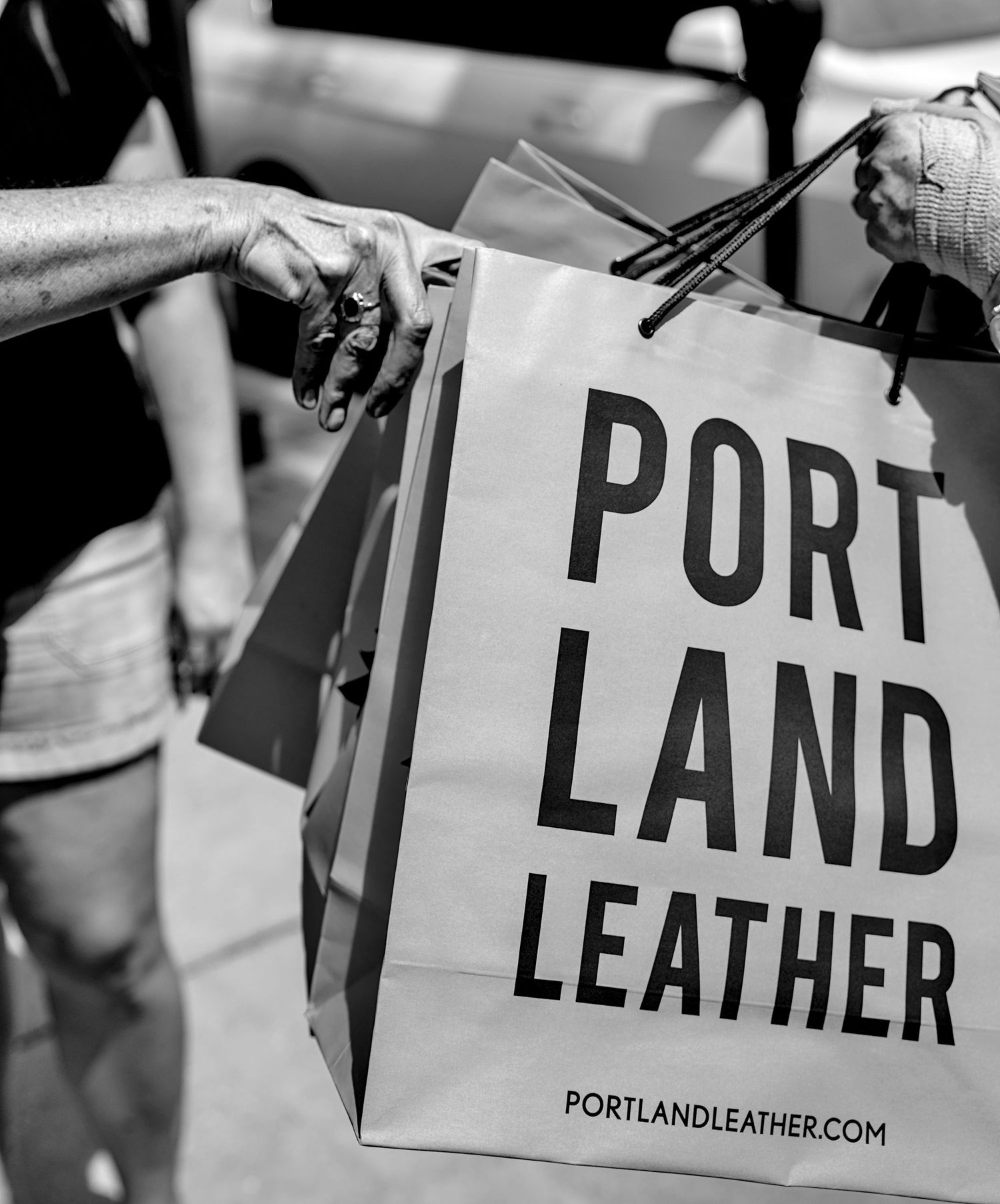 Black and white photo of hands holding a Portland Leather shopping bag outdoors.