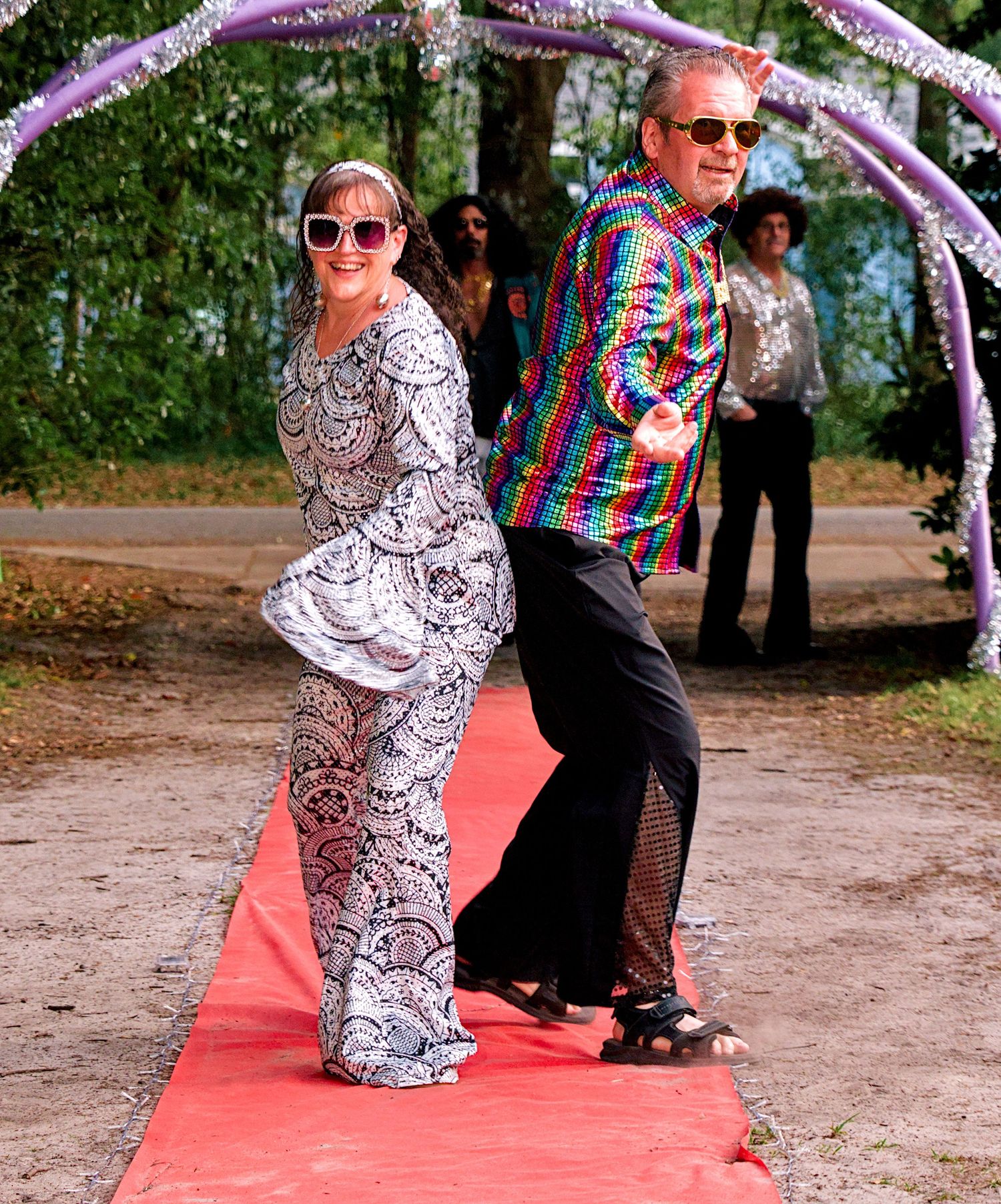 Couple in colorful 70s disco costumes walk red carpet, woman in lace jumpsuit, man in rainbow sequin shirt.