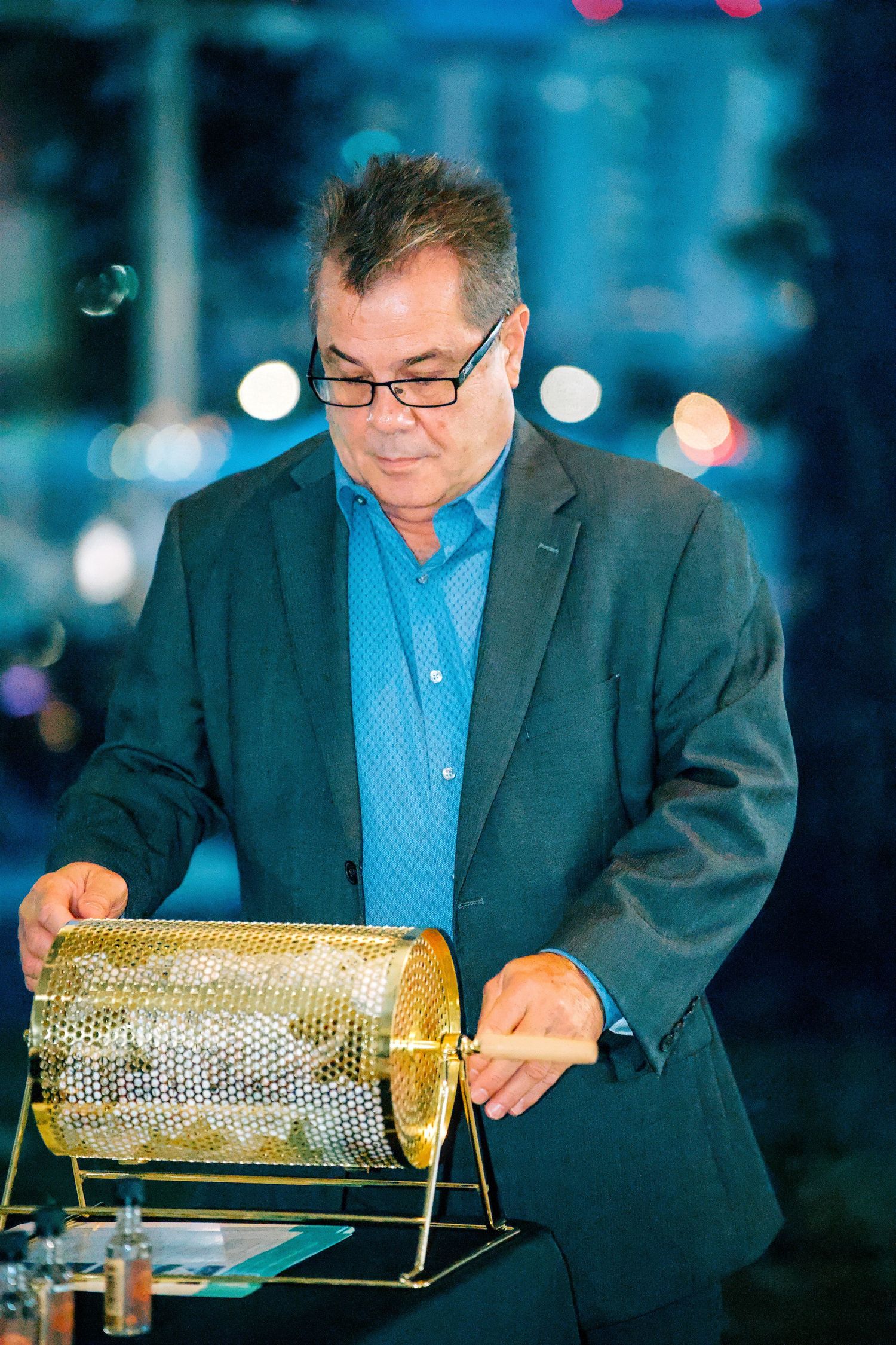 Man in blazer spinning a golden raffle drum at an evening event with bokeh lights in background.