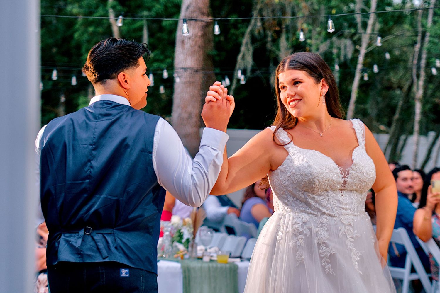 Bride in lace gown shares first dance with partner under string lights at outdoor wedding.
