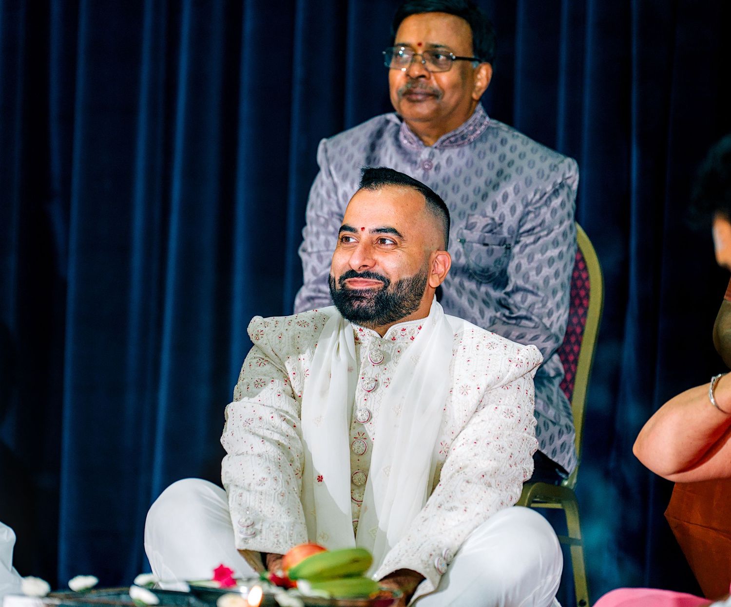 Groom in white sherwani seated during traditional Hindu wedding ceremony with elder behind.
